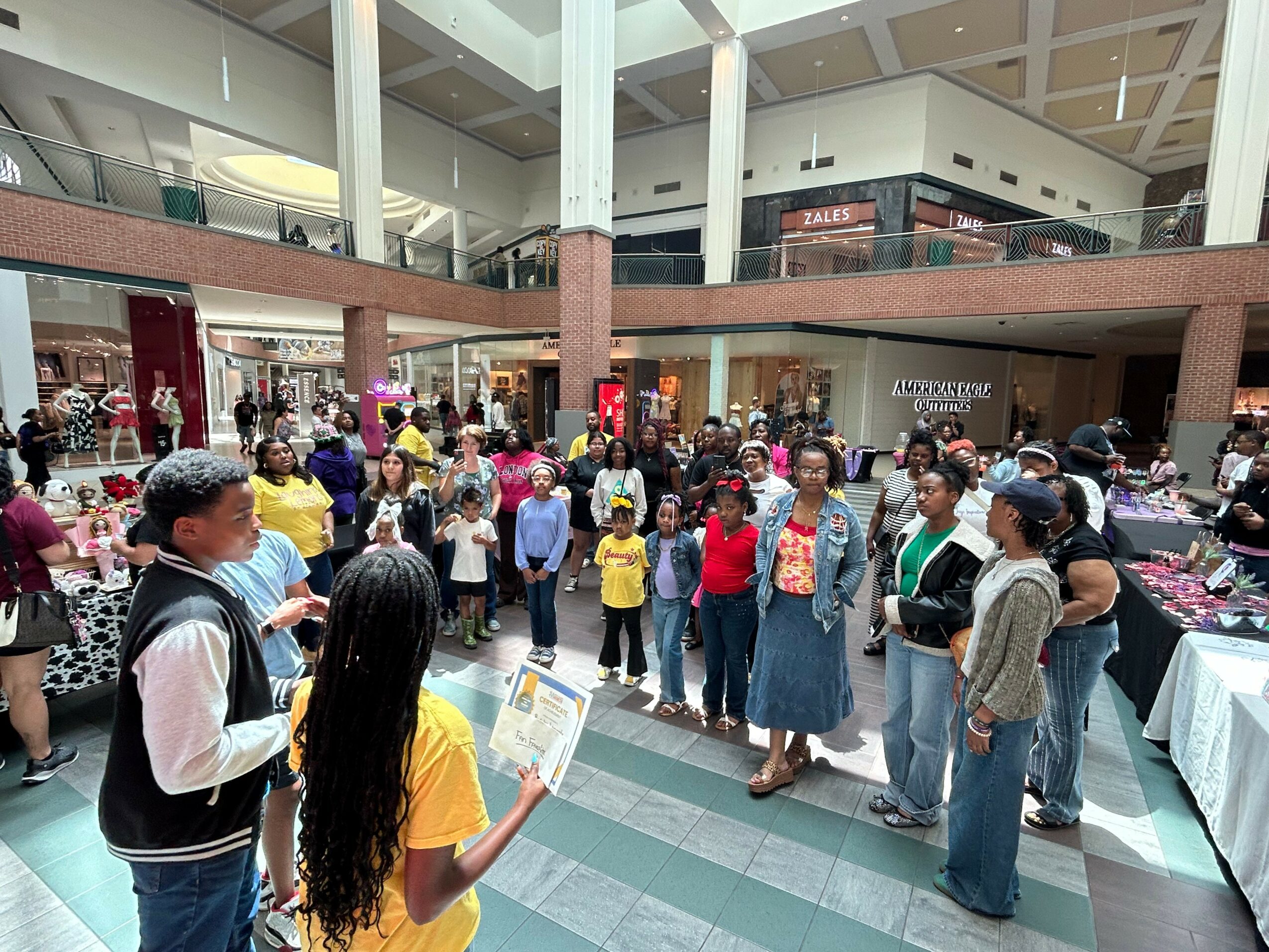 Group of people gathered in a shopping mall, some holding papers, with tables and displays nearby.