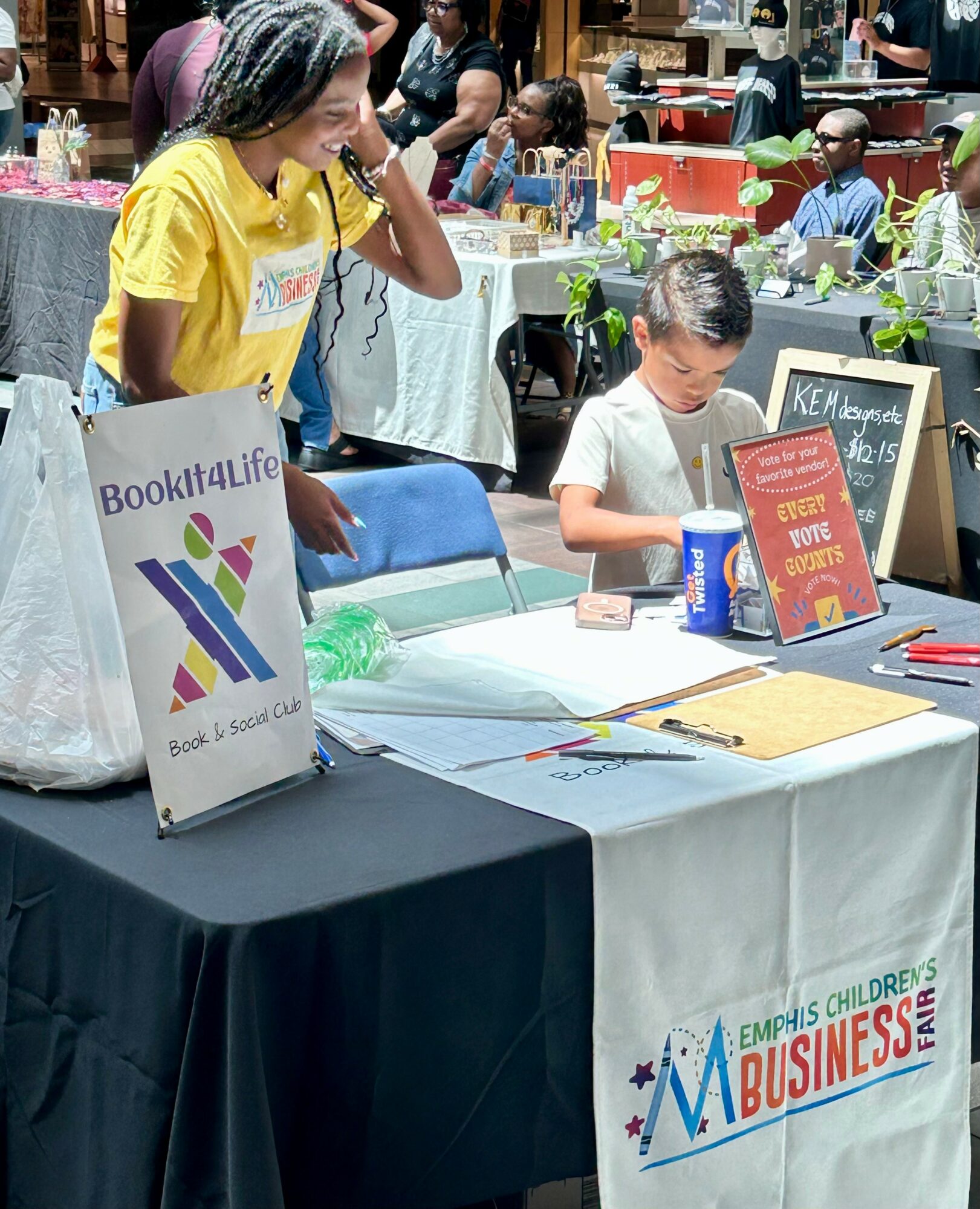 Young girl and boy at a table with books and signs, in a busy indoor event space.