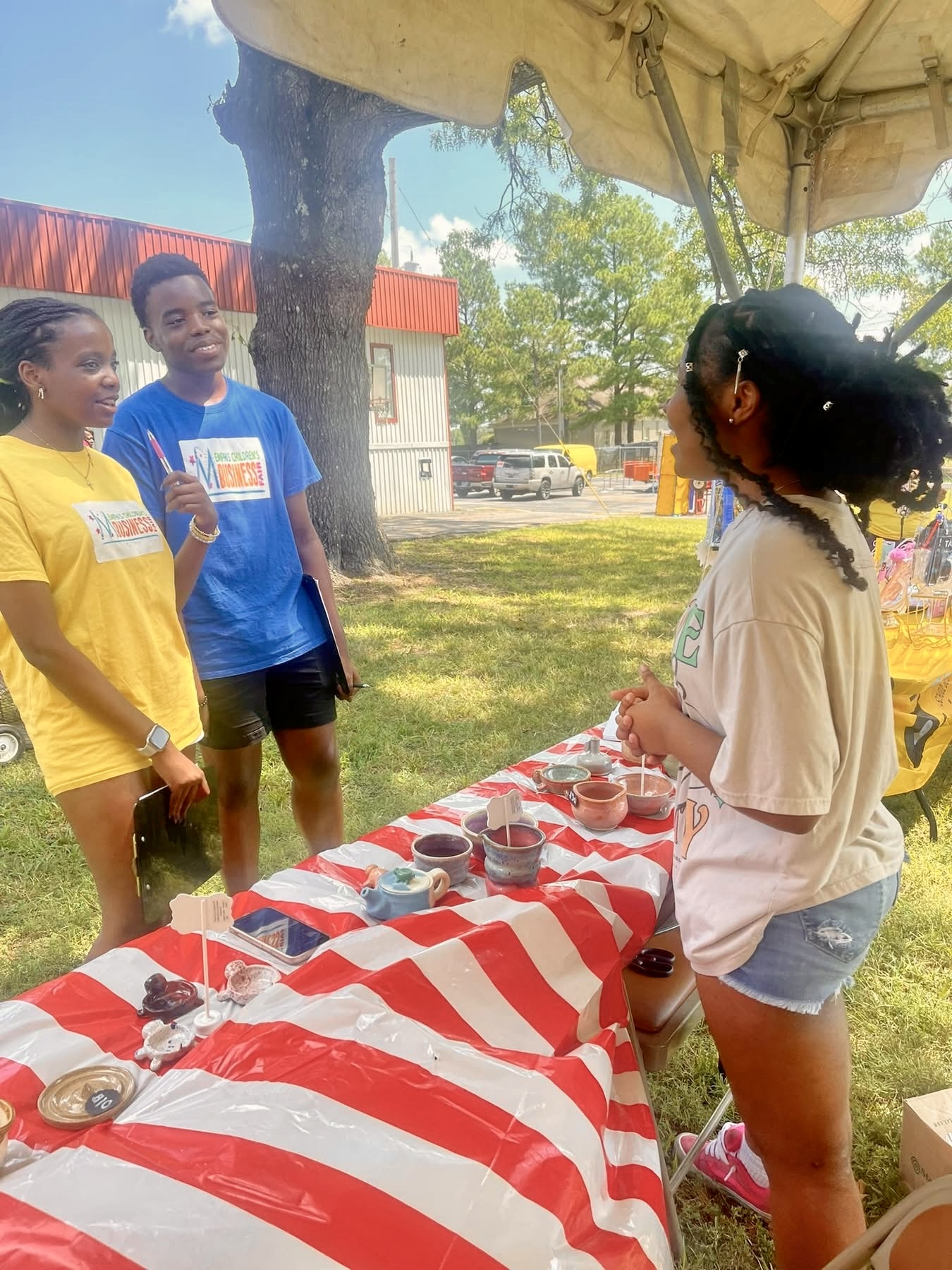 Three people standing outdoors near a table with red and white striped tablecloth, talking to a woman behind the table.