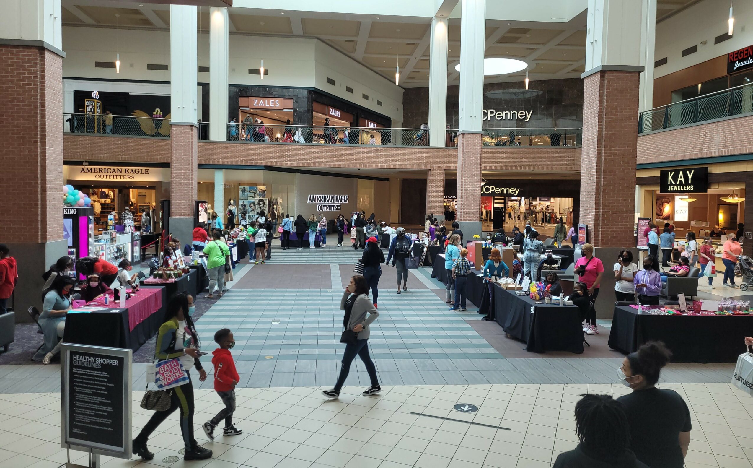 Shopping mall interior with people walking and browsing stores, tables, and kiosks in open central area.