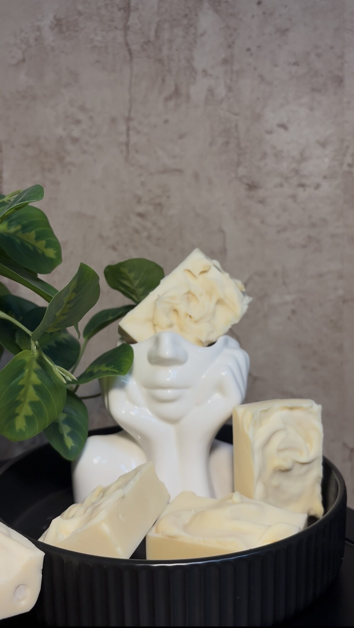 White ceramic vase with a face, filled with white flowers, surrounded by soap bars on a black tray.
