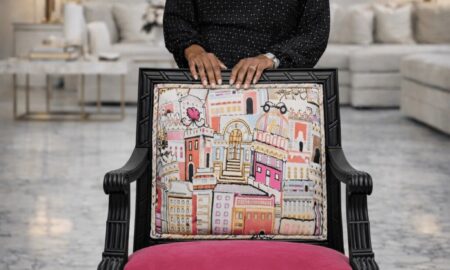 Smiling woman standing behind a decorated chair with a cityscape design, in a modern living room with sofas and lamps.