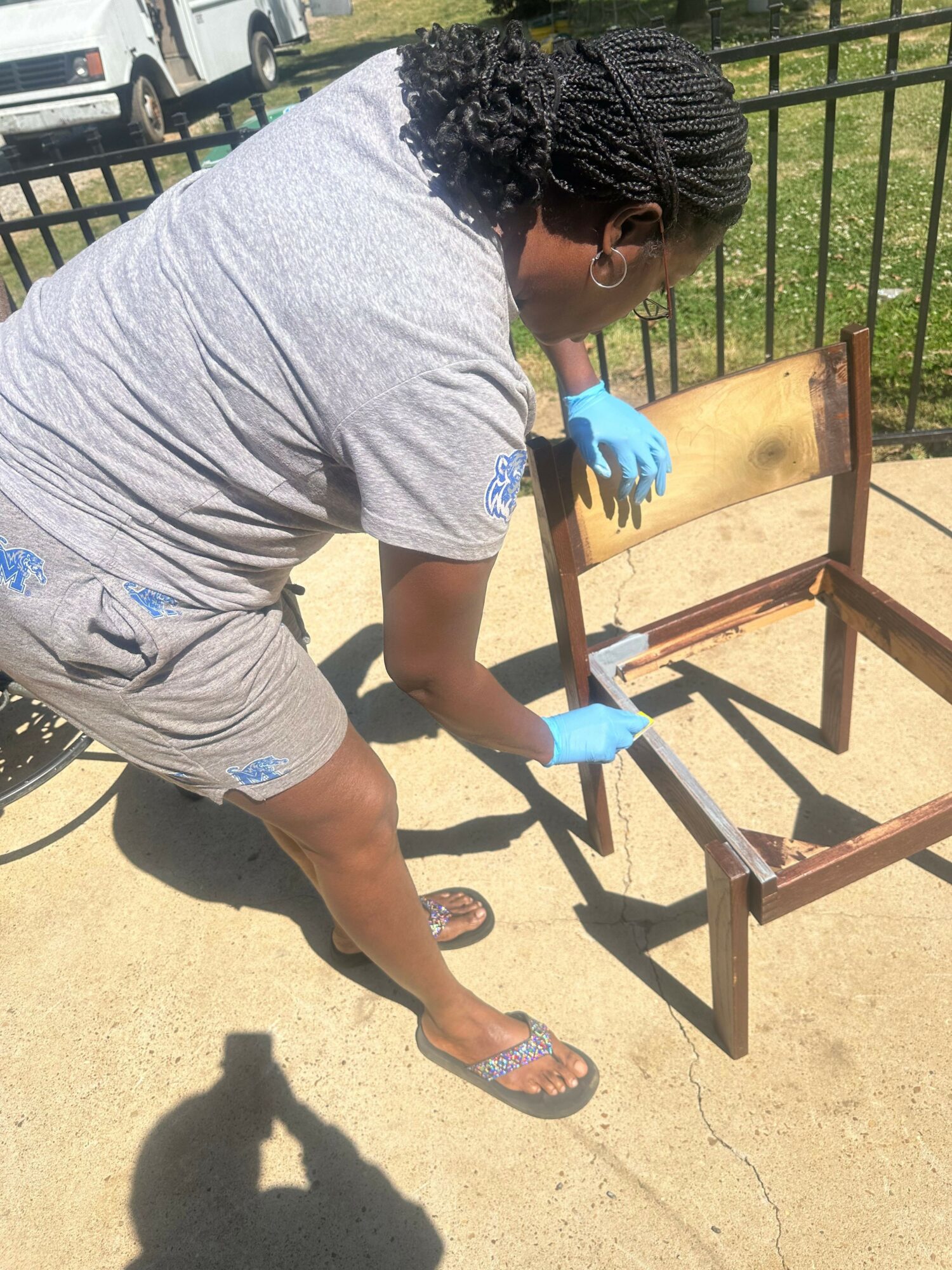 Person wearing gray shirt and shorts, cleaning wooden chair outdoors with blue gloves.