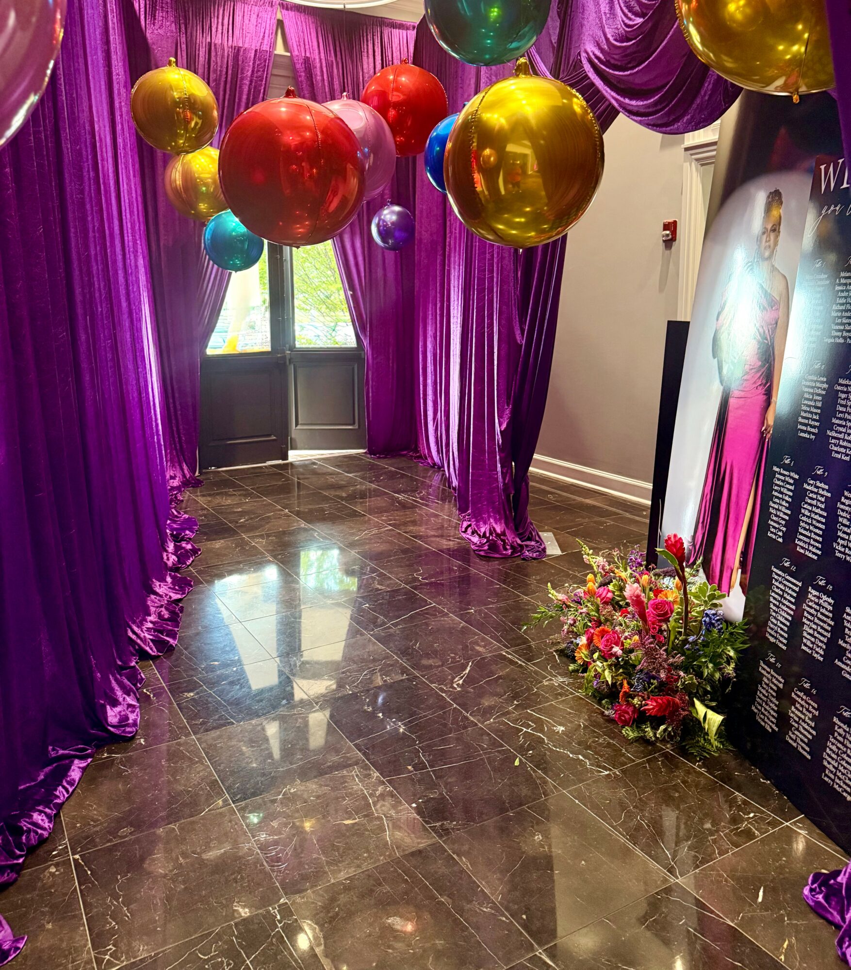 Decorated room with purple curtains, colorful balloons, and a floral arrangement near a display board.