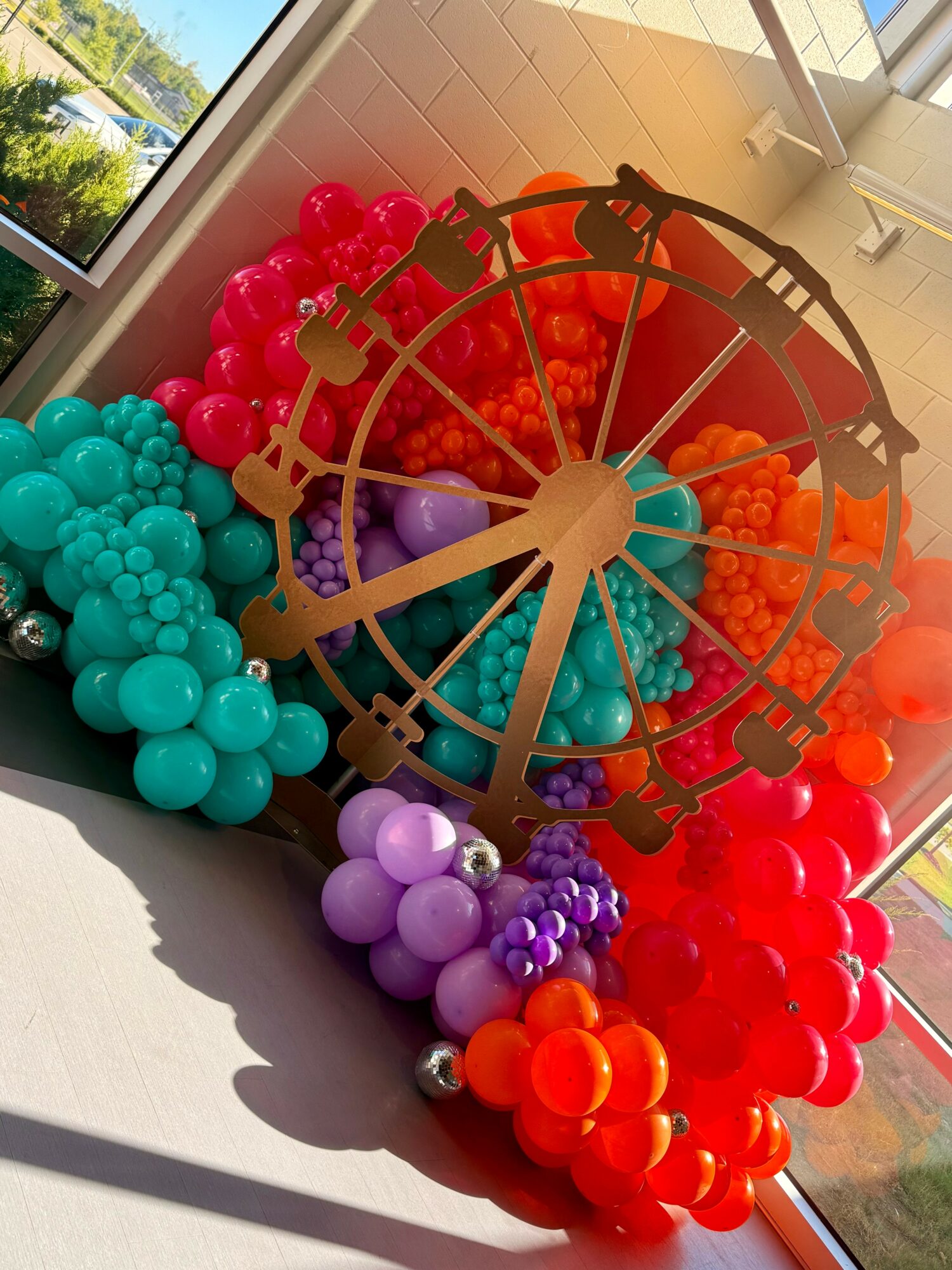 Colorful balloons arranged around a clock face with gold hands, placed near a window with sunlight and outdoor view.