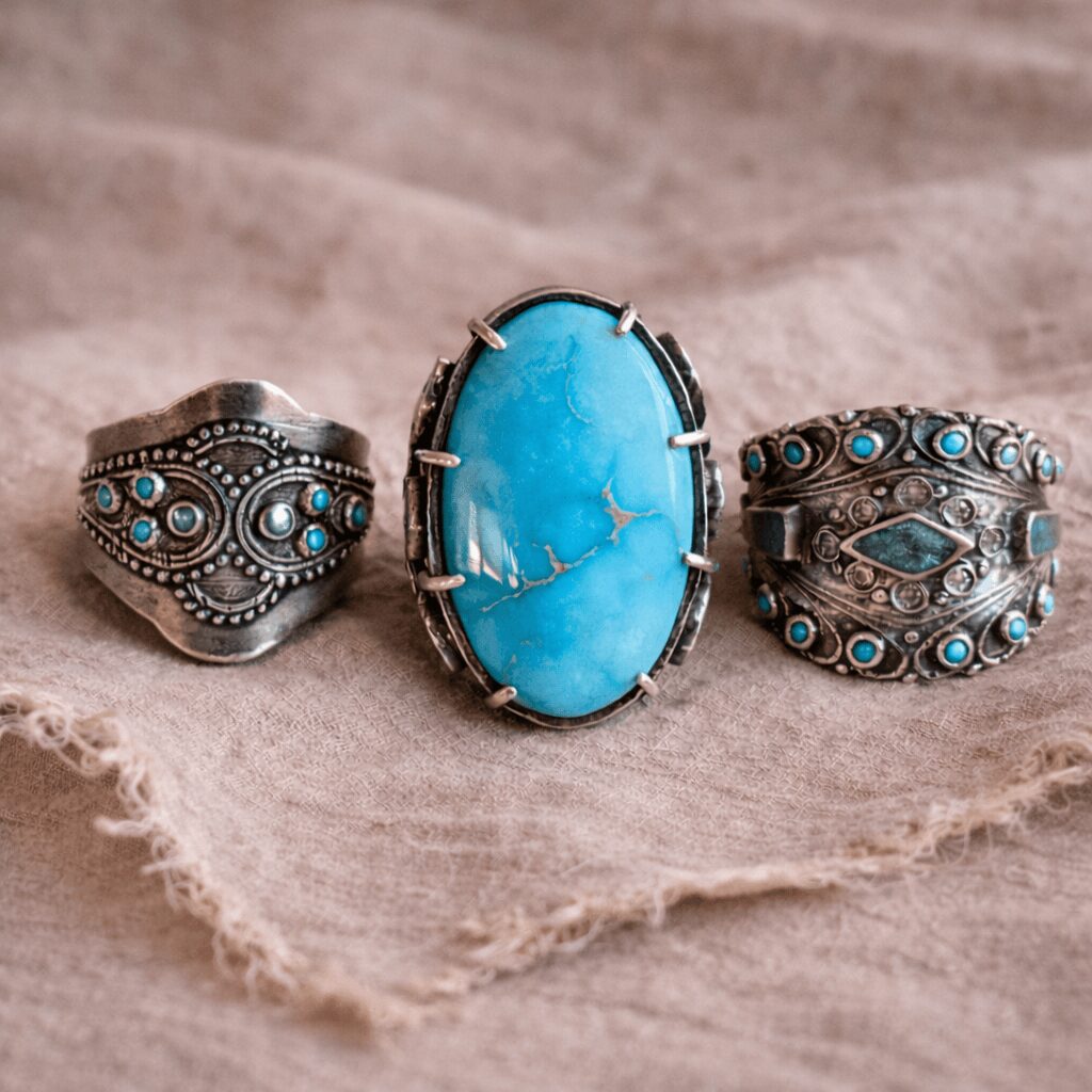 Three silver rings with turquoise stones and intricate designs, displayed on a textured fabric surface.