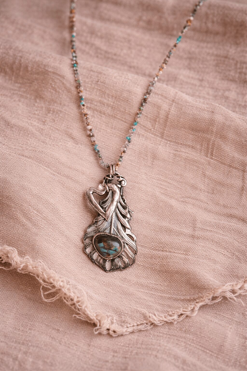 Silver necklace with a large leaf-shaped pendant and a small gemstone, resting on pink fabric with frayed edges.