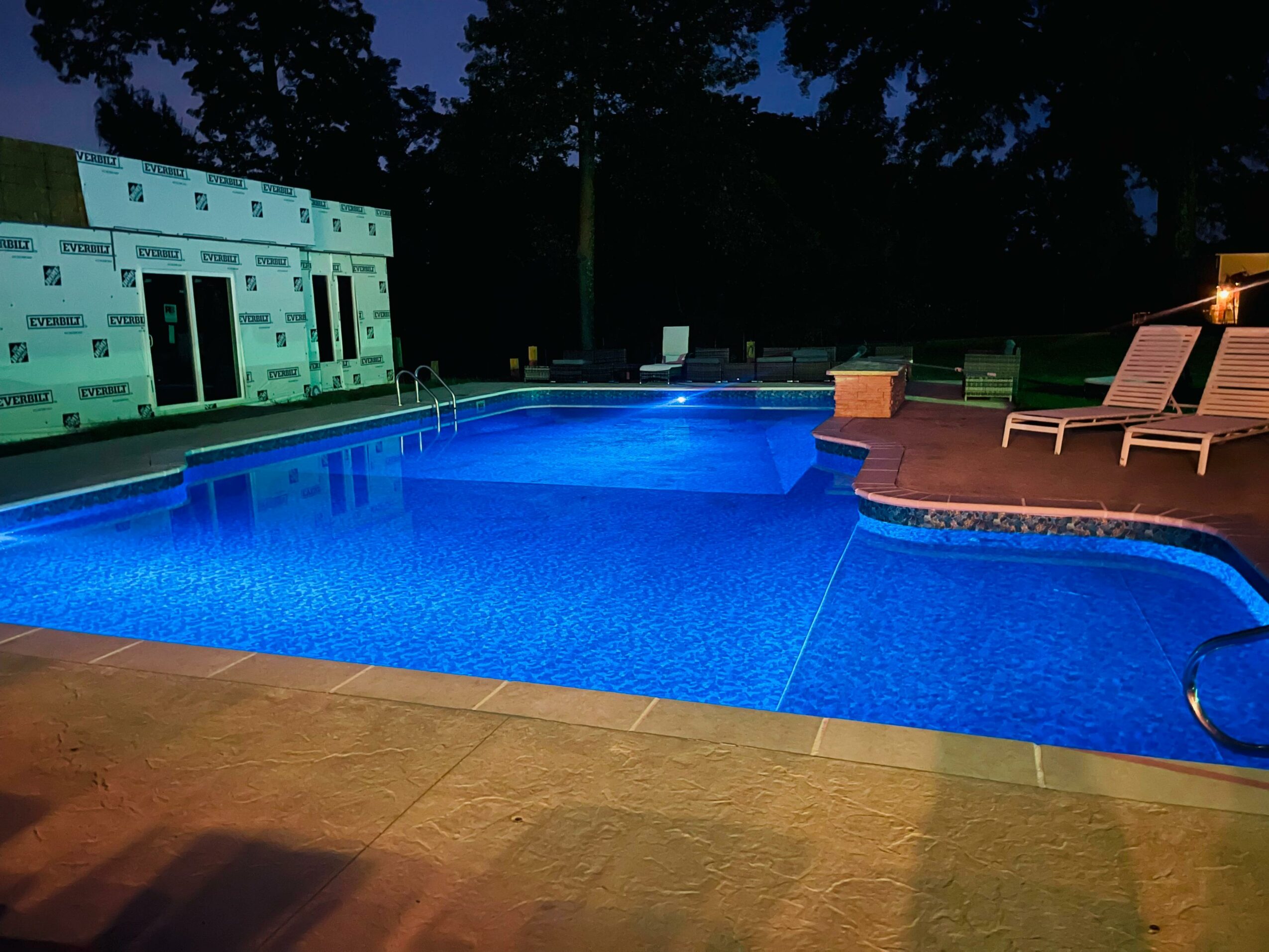 Swimming pool illuminated at night with lounge chairs on the deck, surrounded by trees and a building under construction.