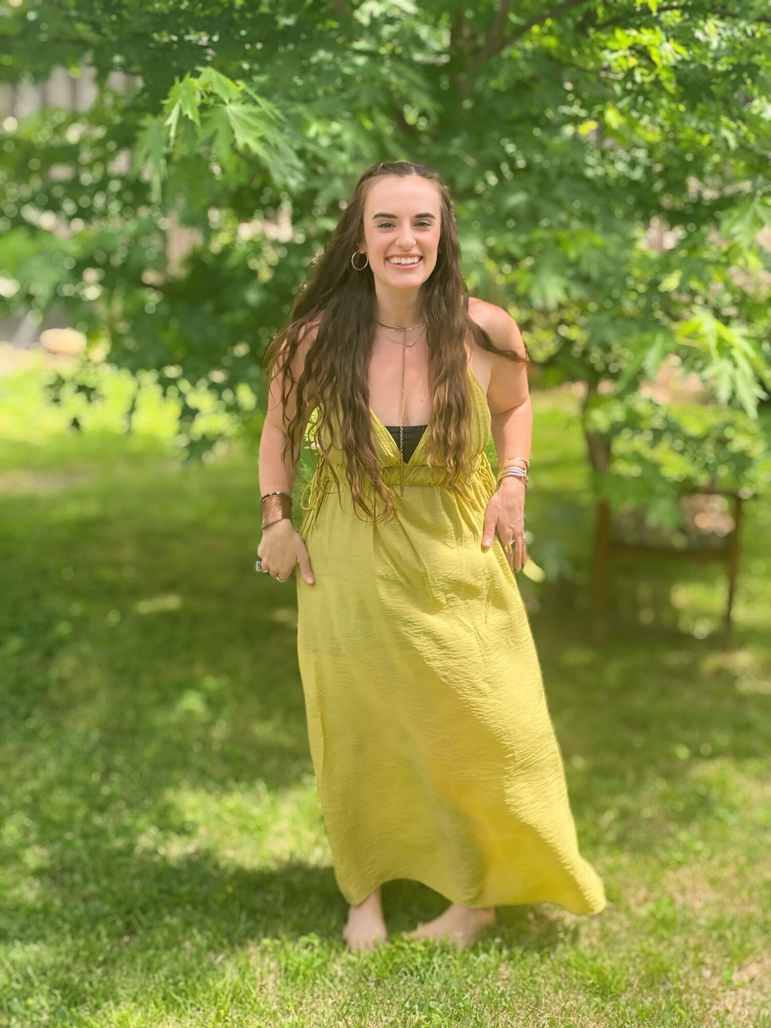 Young woman with long hair in a yellow dress standing on grass with green trees in background.