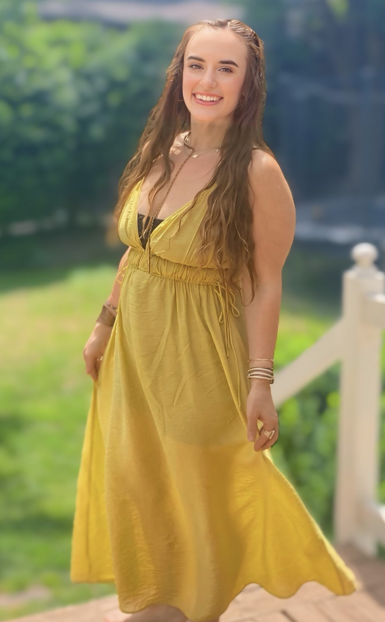 Young woman with long hair in a yellow dress outdoors, smiling, with greenery and a white fence in background.
