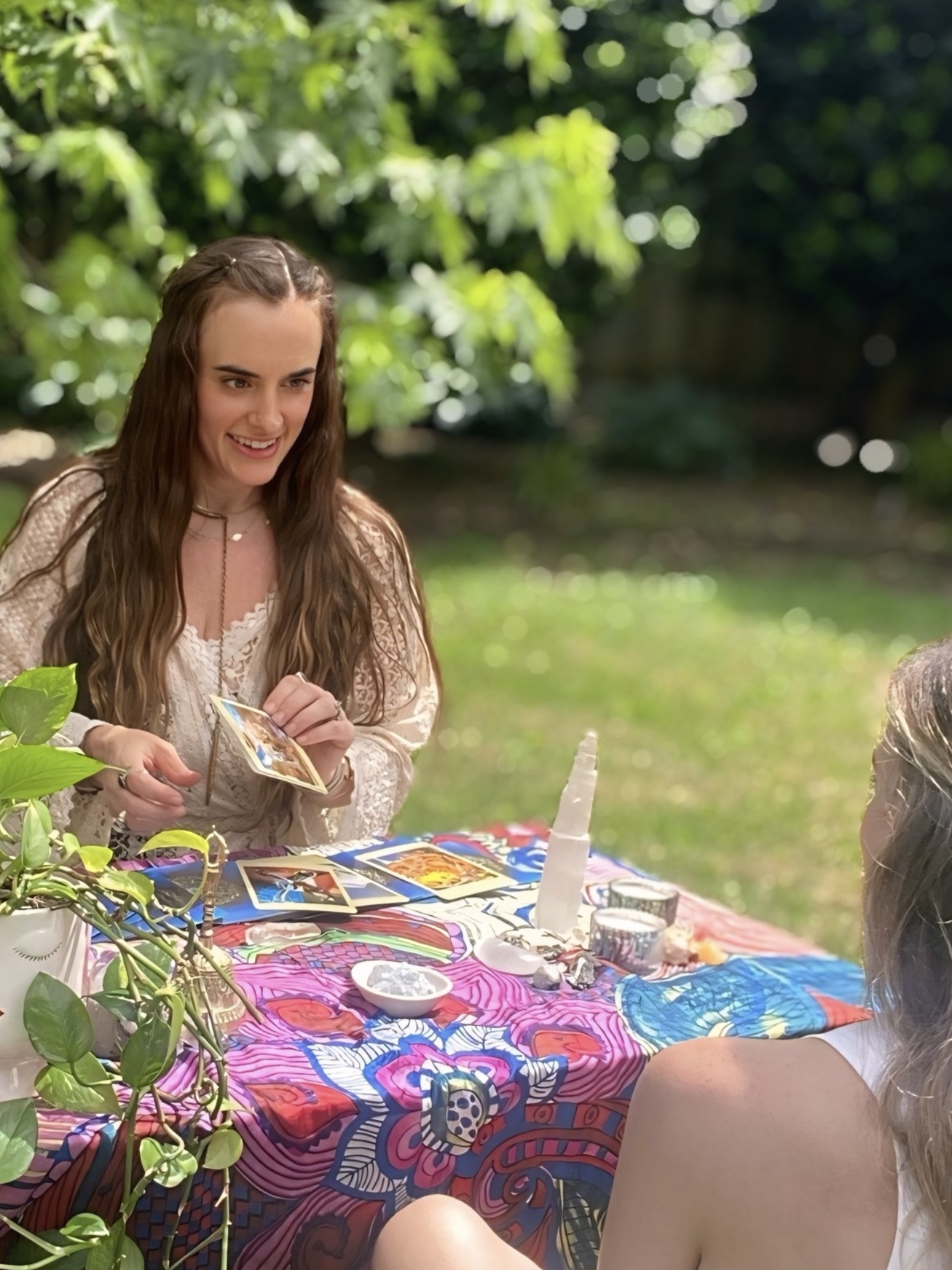 Young woman with long brown hair smiling, sitting at a colorful table outdoors with a person whose back is visible.