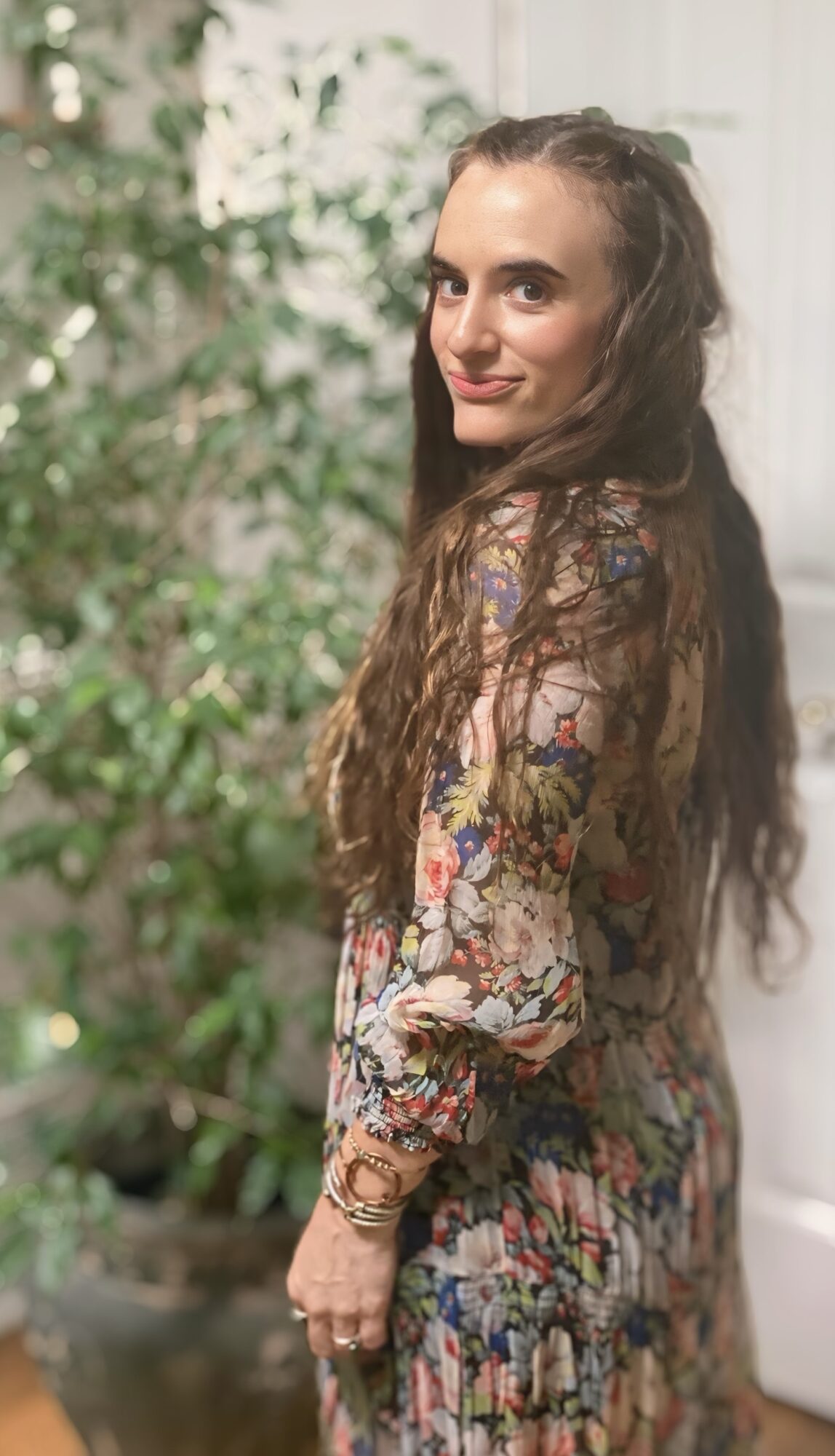 Young woman with long wavy hair wearing a floral dress, standing outdoors with greenery in background.