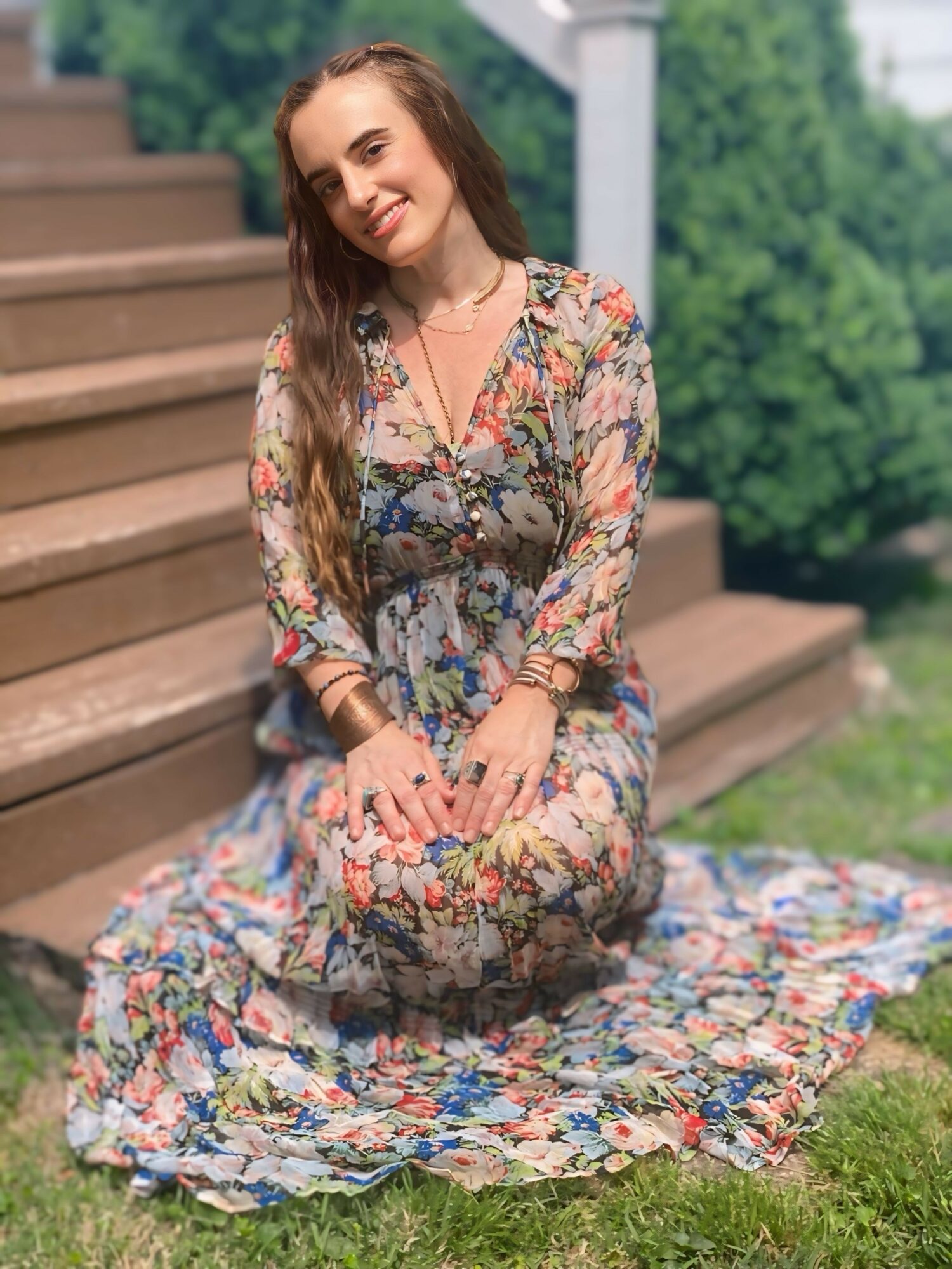 Young woman kneeling on grass in floral dress, smiling outdoors near stairs and greenery.