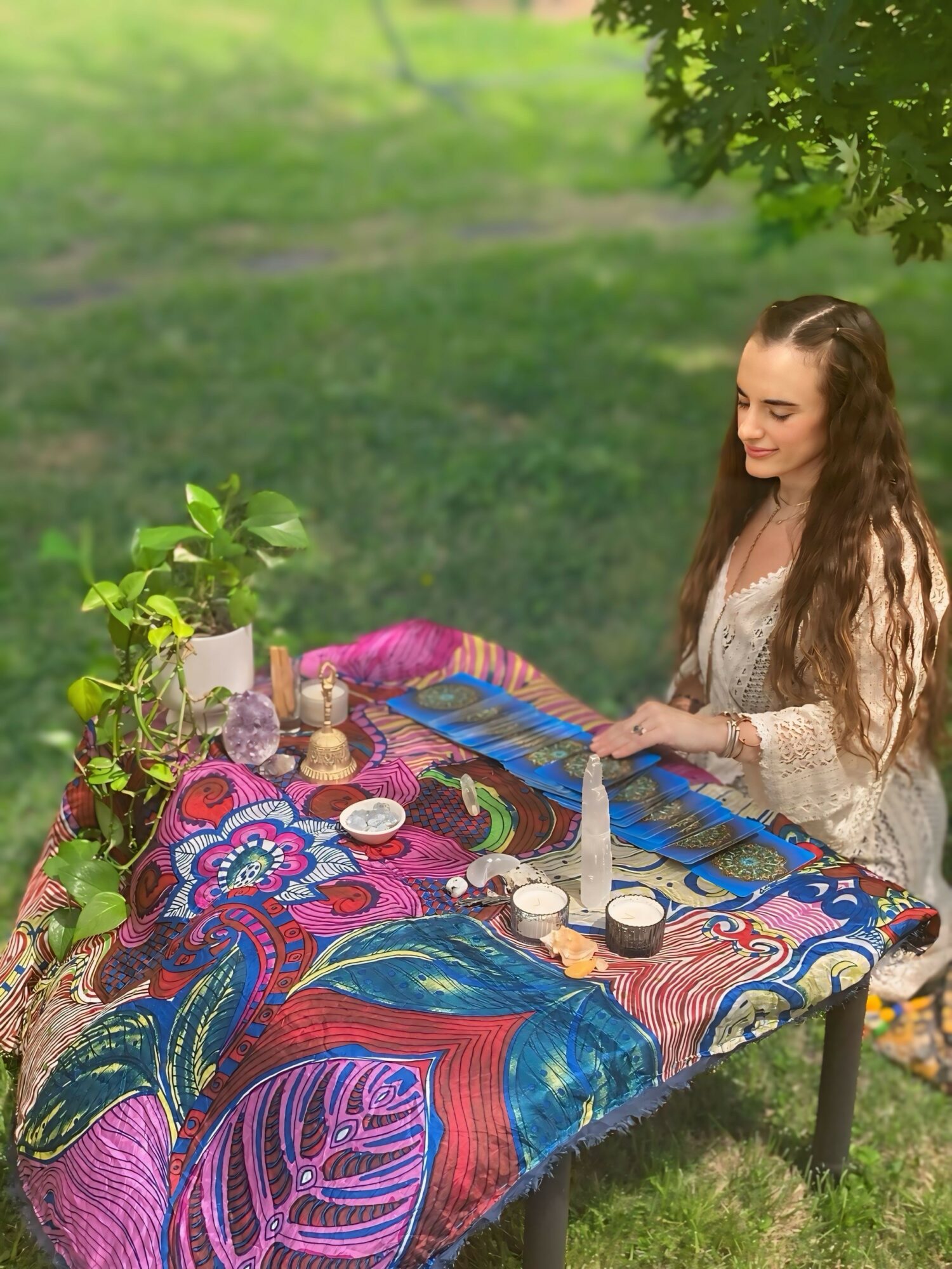 Young woman with long hair sitting at a colorful table outdoors, surrounded by plants and decorative items.