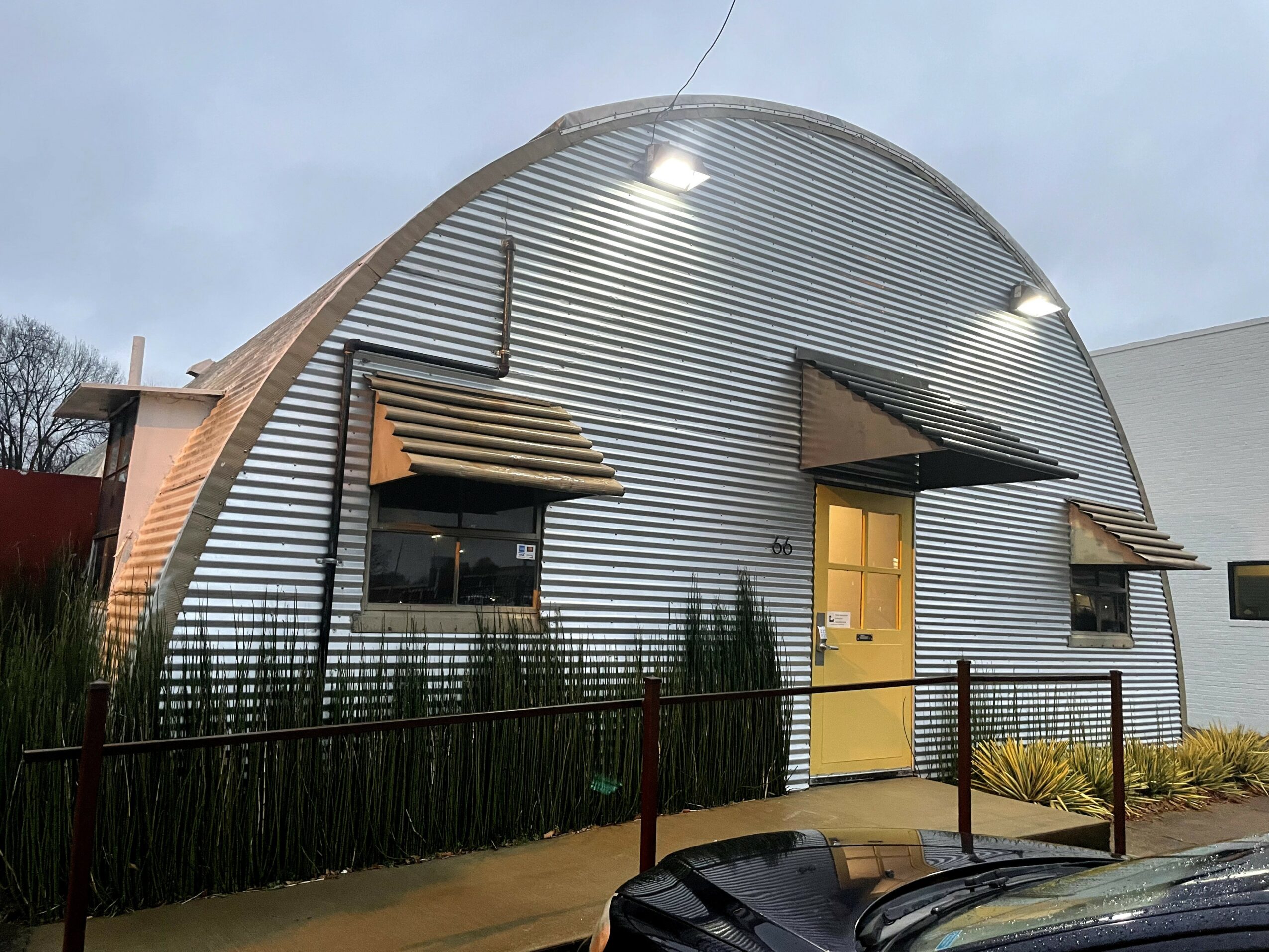 A curved metal building with three small awnings over windows and a yellow door, surrounded by a fence and plants.