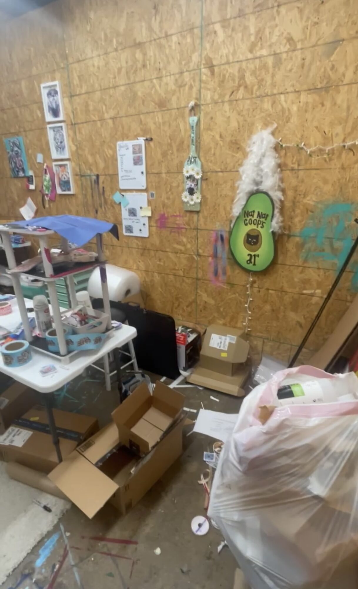 Cluttered room with boxes, a table, and various items against a wooden wall with posters and decorations.