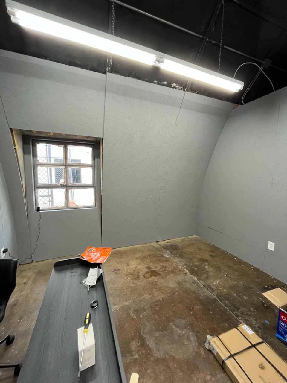 Empty room with a window, unfinished floor, and a table with items, including a box and a plastic bag.