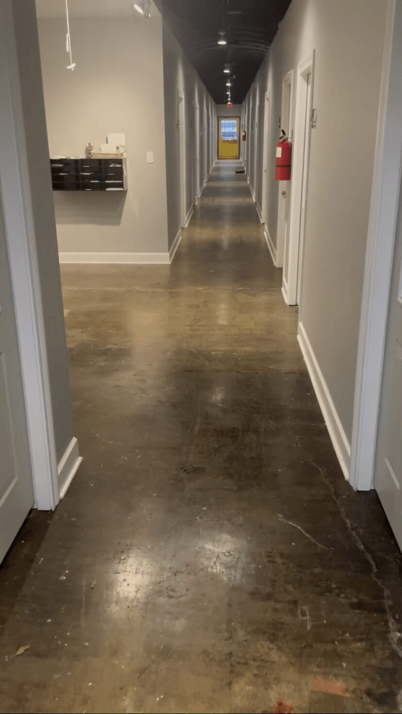 Long corridor with wooden floor, white walls, doors on both sides, and a fire extinguisher on the right wall.