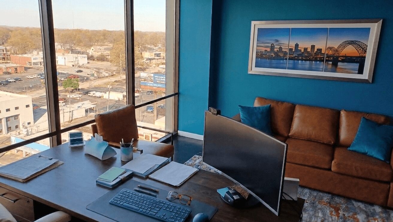 Office desk with computer monitor, keyboard, mouse, and documents, near large window with city view, blue wall with framed picture.