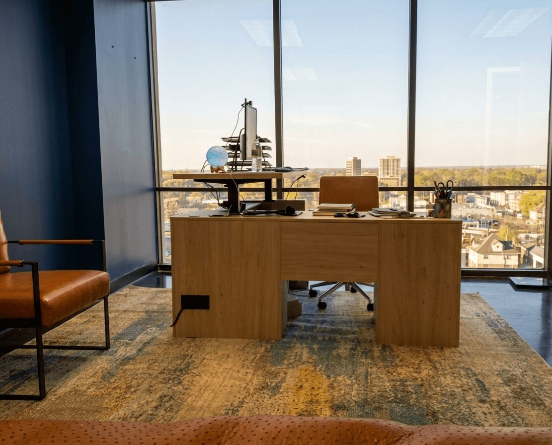 Office desk with a chair, view of cityscape through large window, items on desk, and a brown chair nearby.
