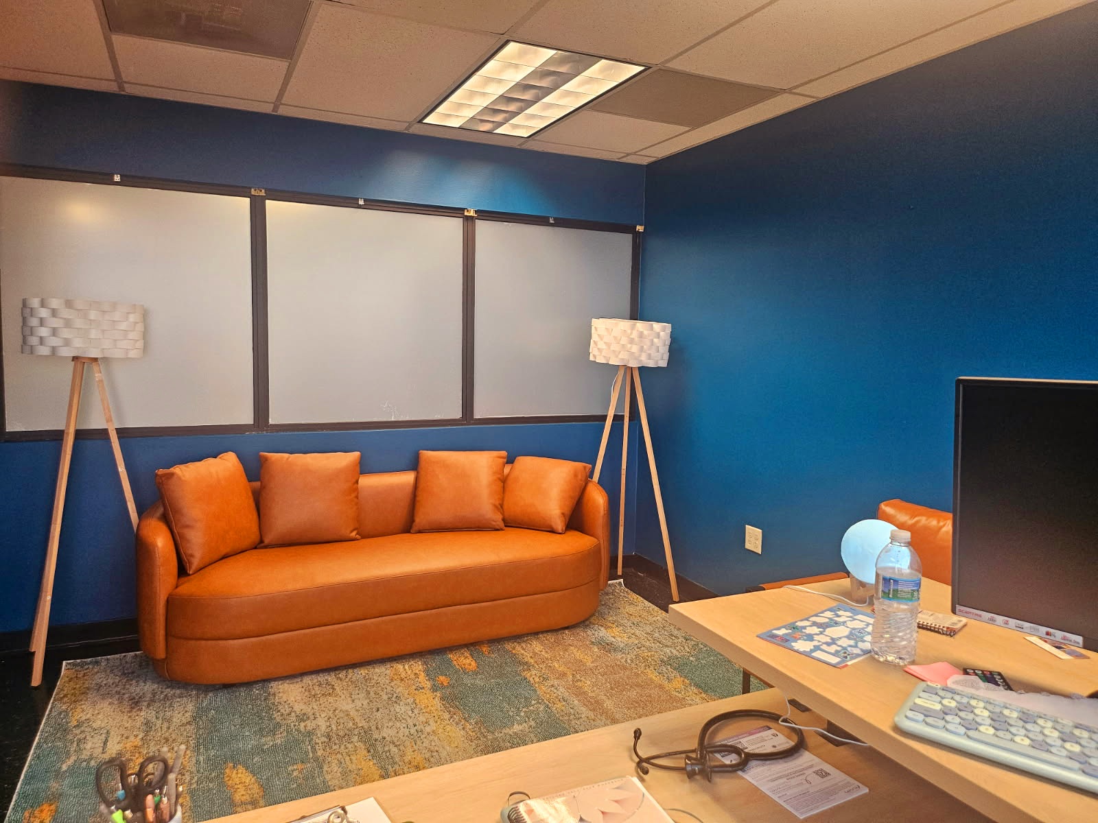 Living room with orange sofa, blue wall, two floor lamps, and a desk with a monitor and water bottle.