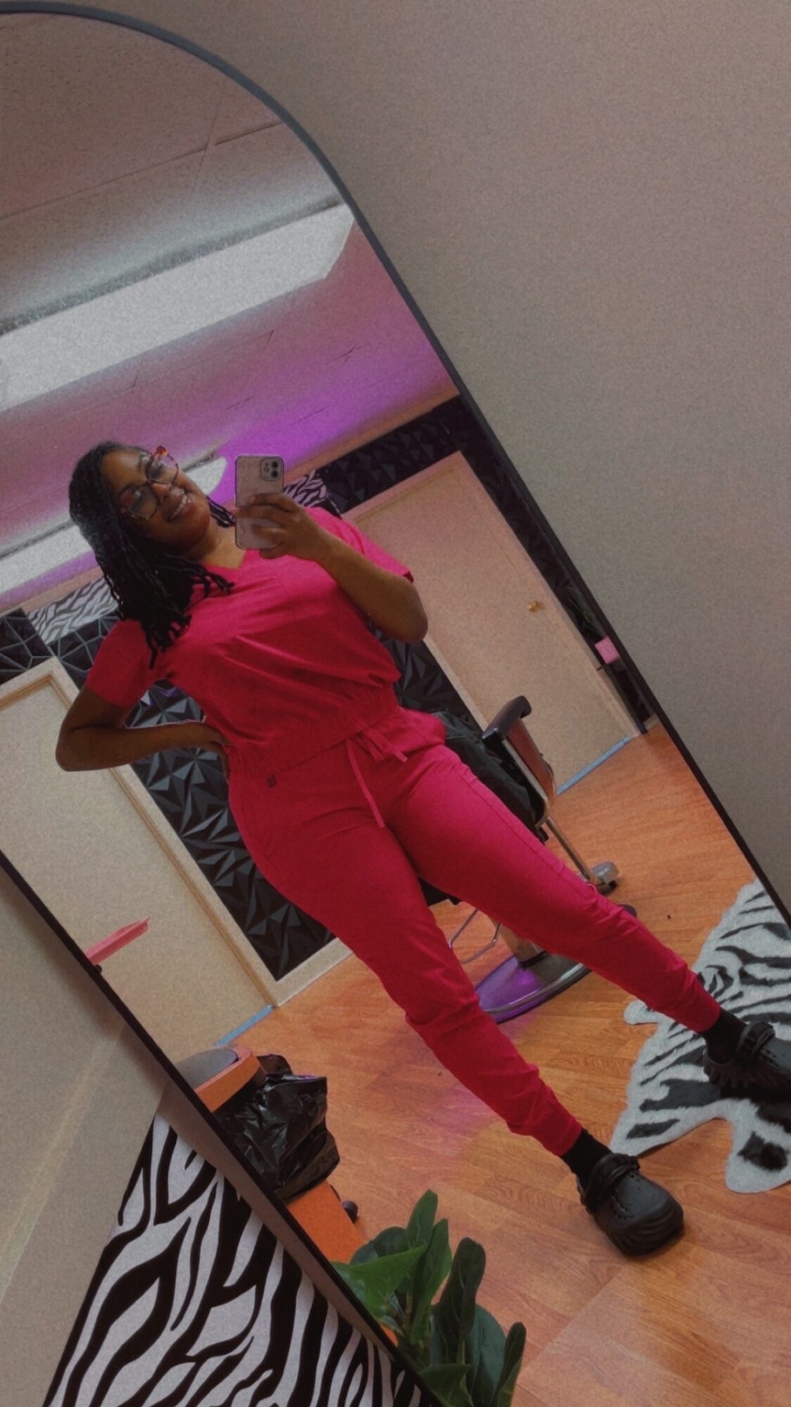 Person taking a mirror selfie in a room with pink and purple lighting, wearing pink outfit and black shoes.