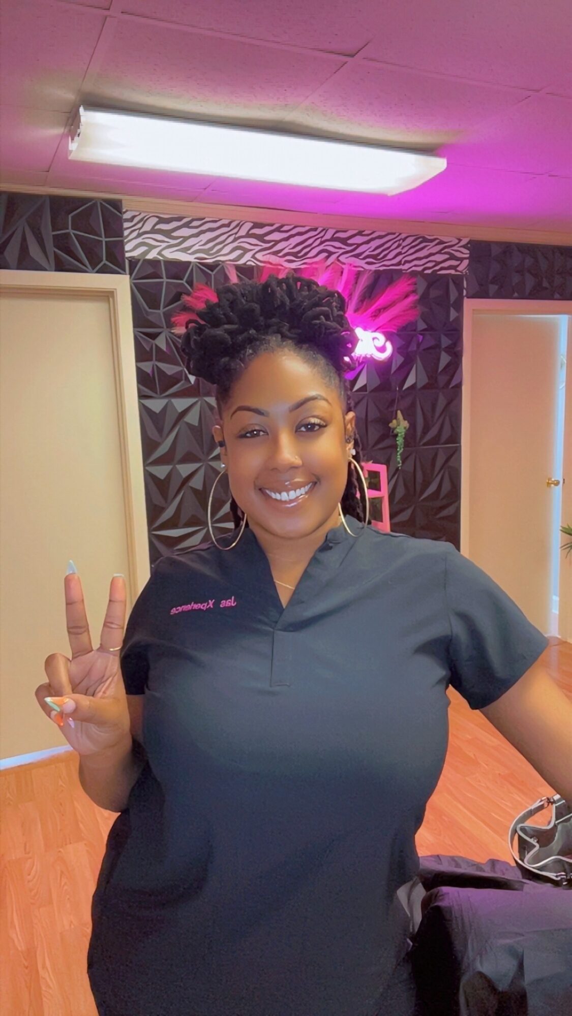 Smiling woman in dark uniform making a peace sign indoors with pink and purple lighting.