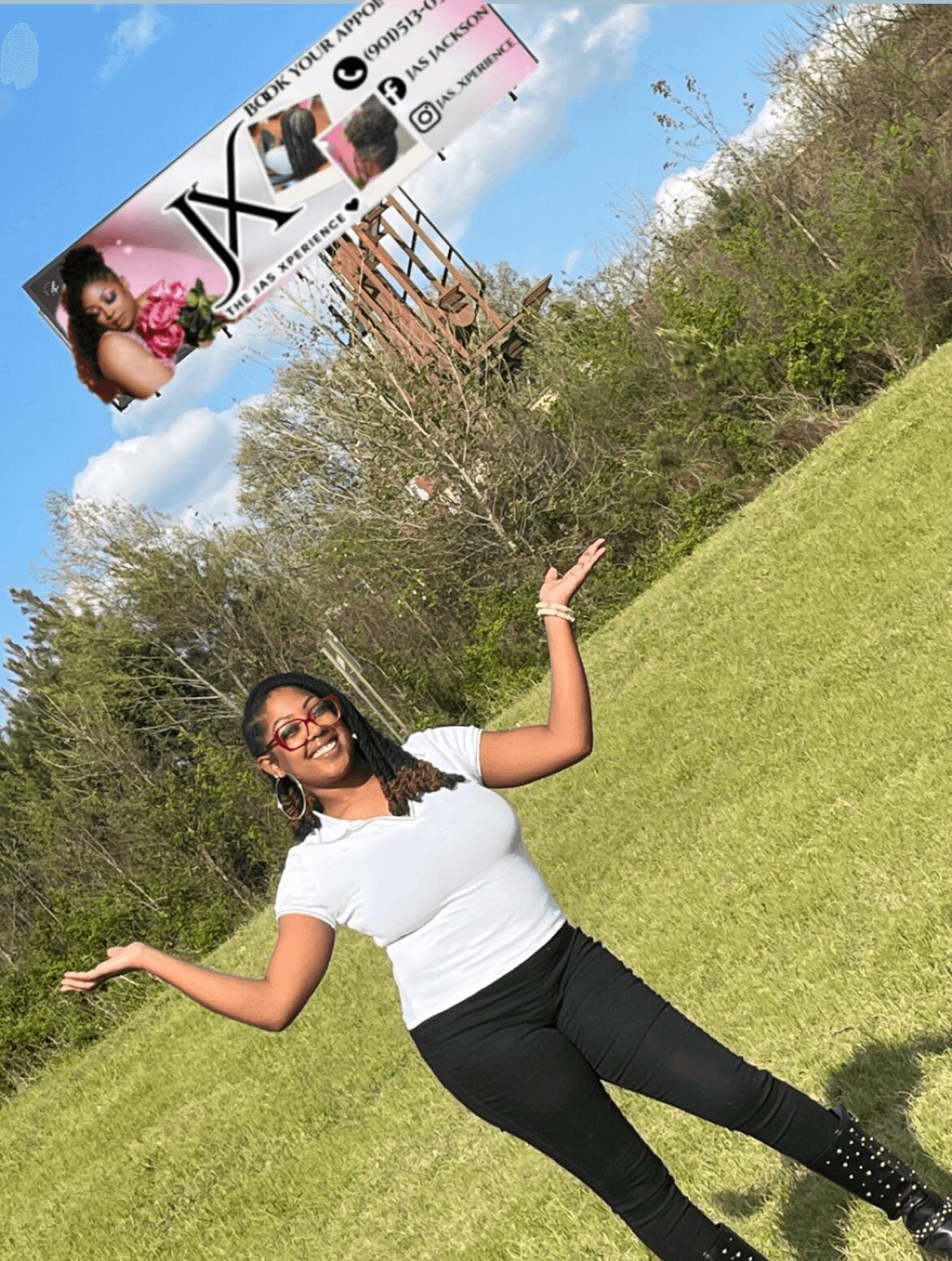 Woman standing on grass, smiling, with arms raised, outdoors with trees and blue sky in background.
