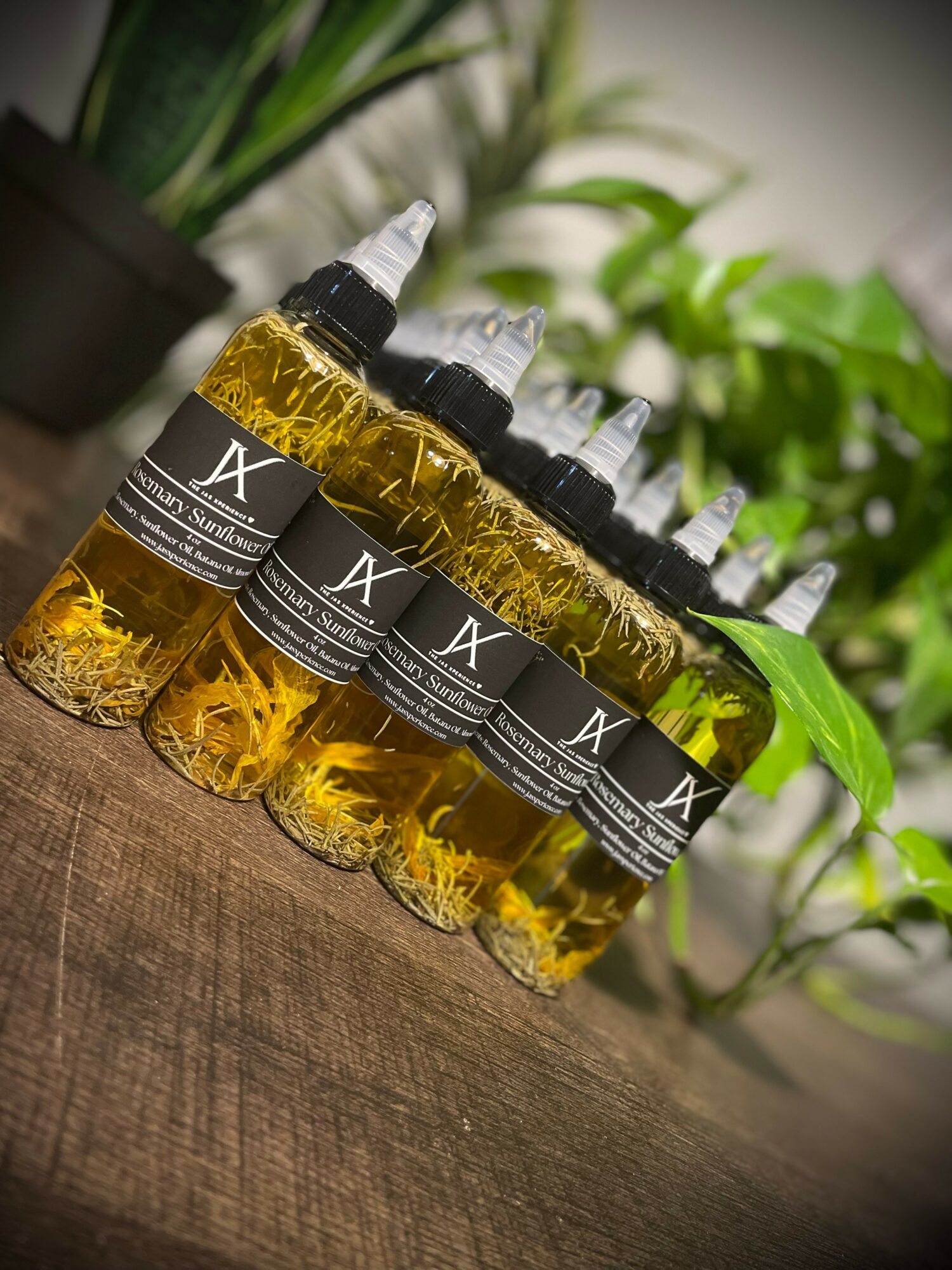 Six bottles with black labels and dropper caps filled with yellow liquid are arranged on a wooden surface with green plants in the background.