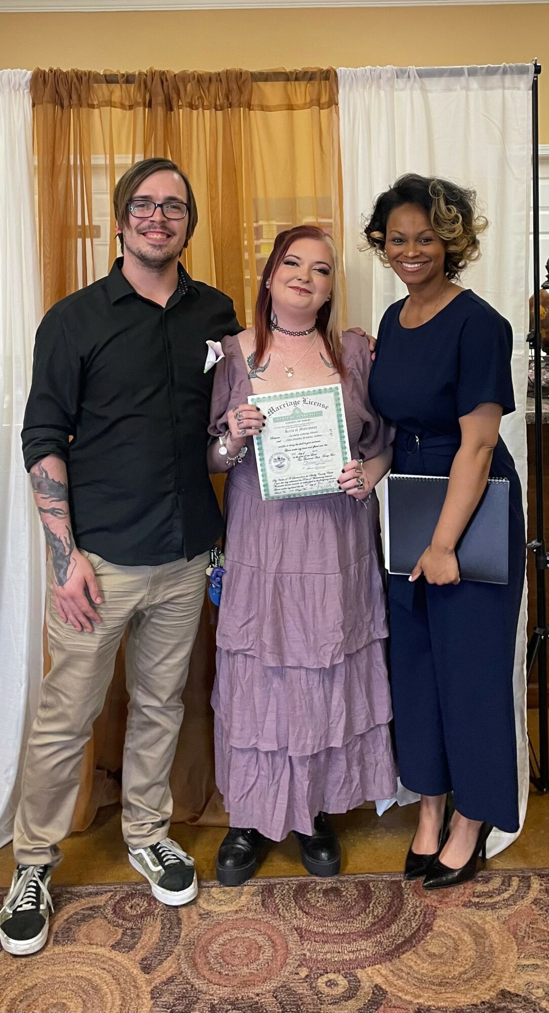 Three people standing indoors, one holding a certificate, smiling, with curtains in the background.