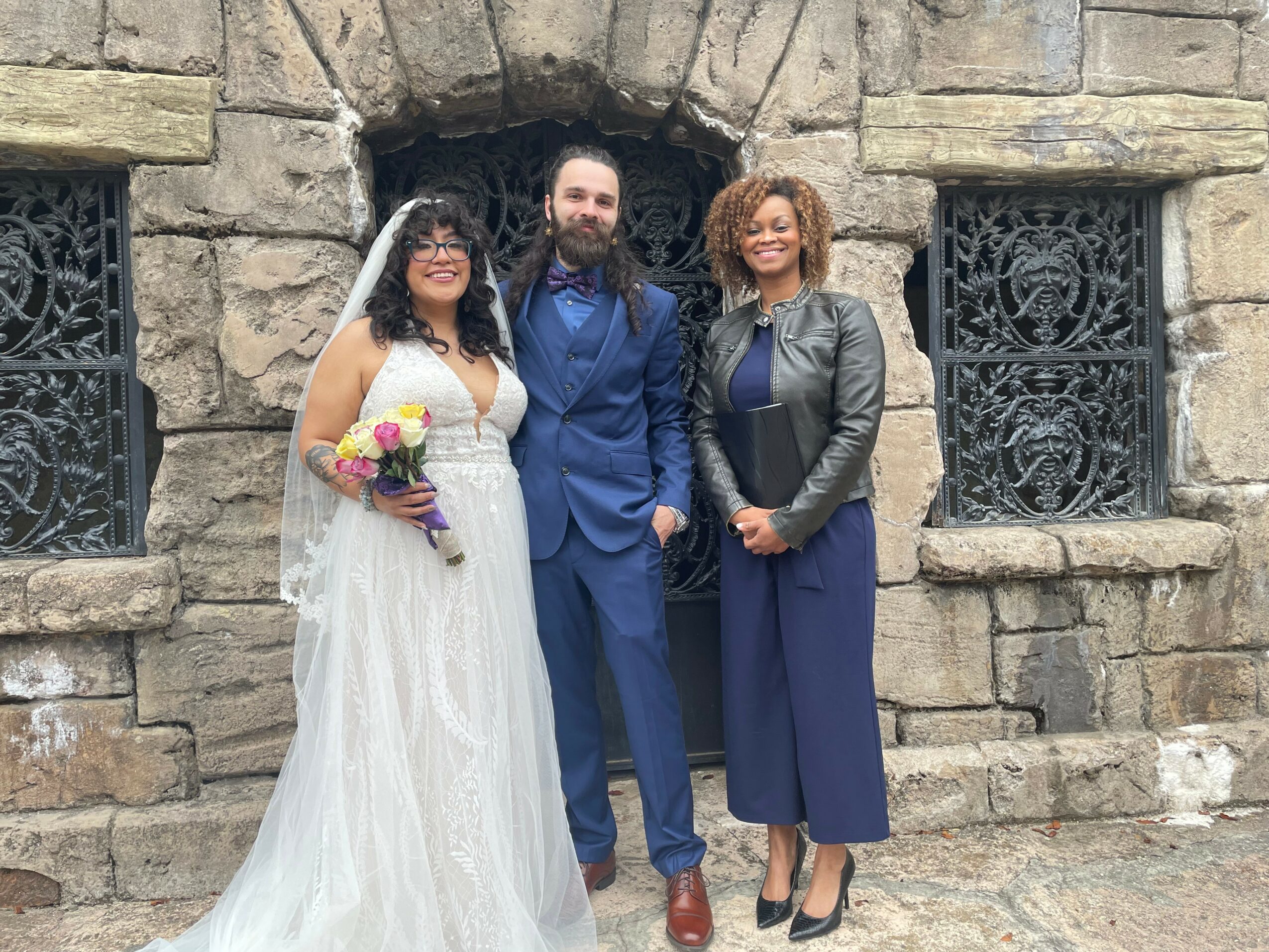 Three people standing in front of a stone wall with decorative windows, one in wedding attire, two in formal clothing.