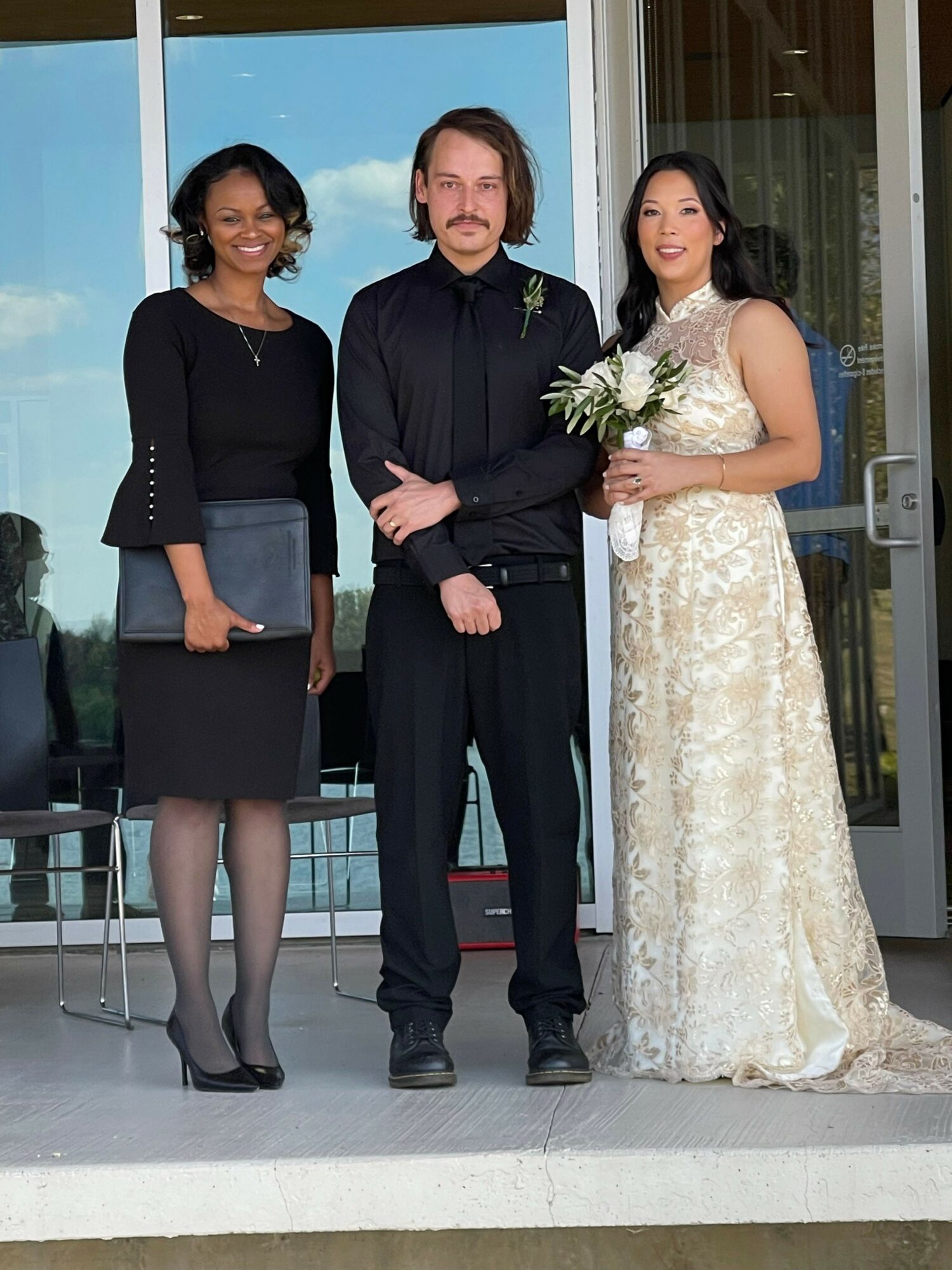 Three people standing outside a building, one woman holding a bouquet, all dressed formally, smiling.