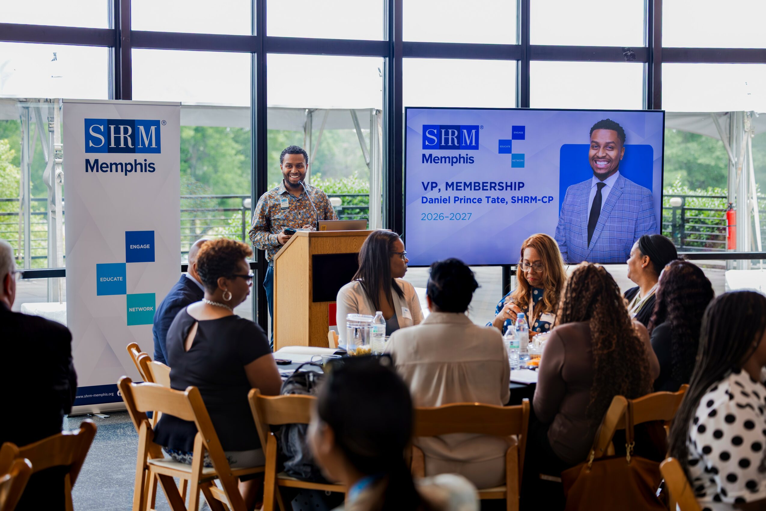 A speaker at a podium addresses an audience in a bright room with large windows, with a large screen displaying a man in a suit.