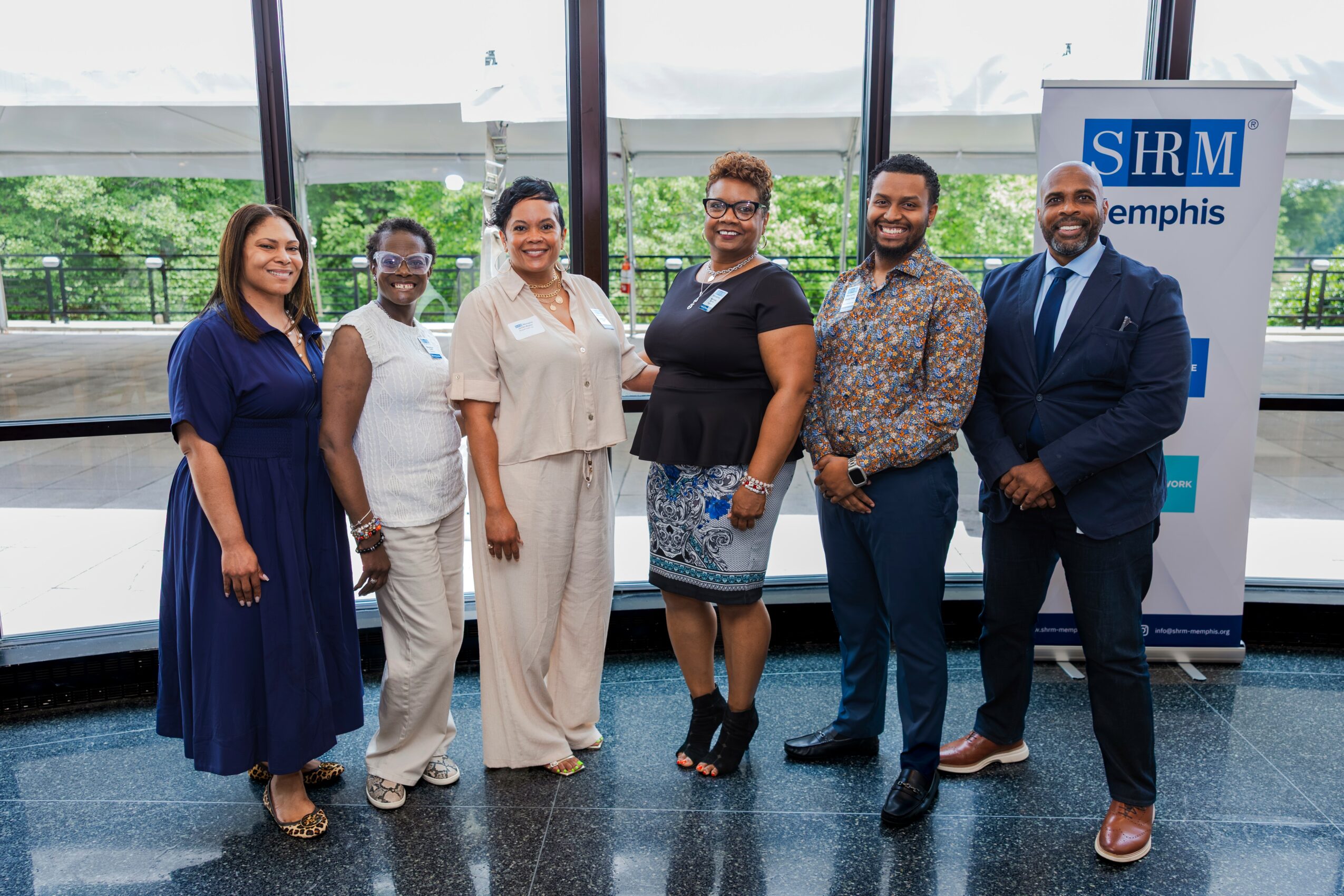 Group of six diverse people standing indoors near windows, smiling, with a SHRM Memphis banner in the background.