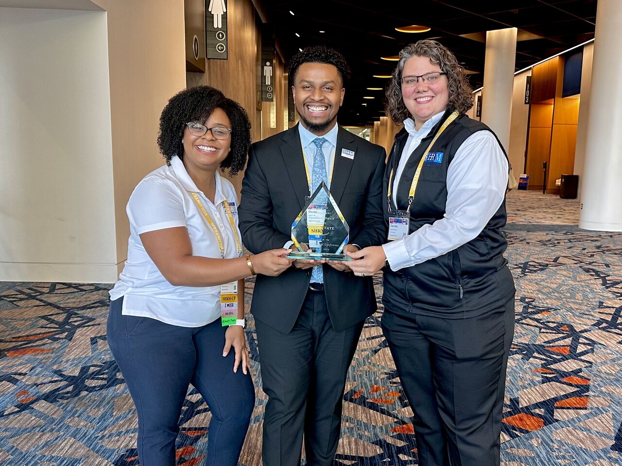 Three people in business attire holding a glass award, smiling in a conference center hallway.