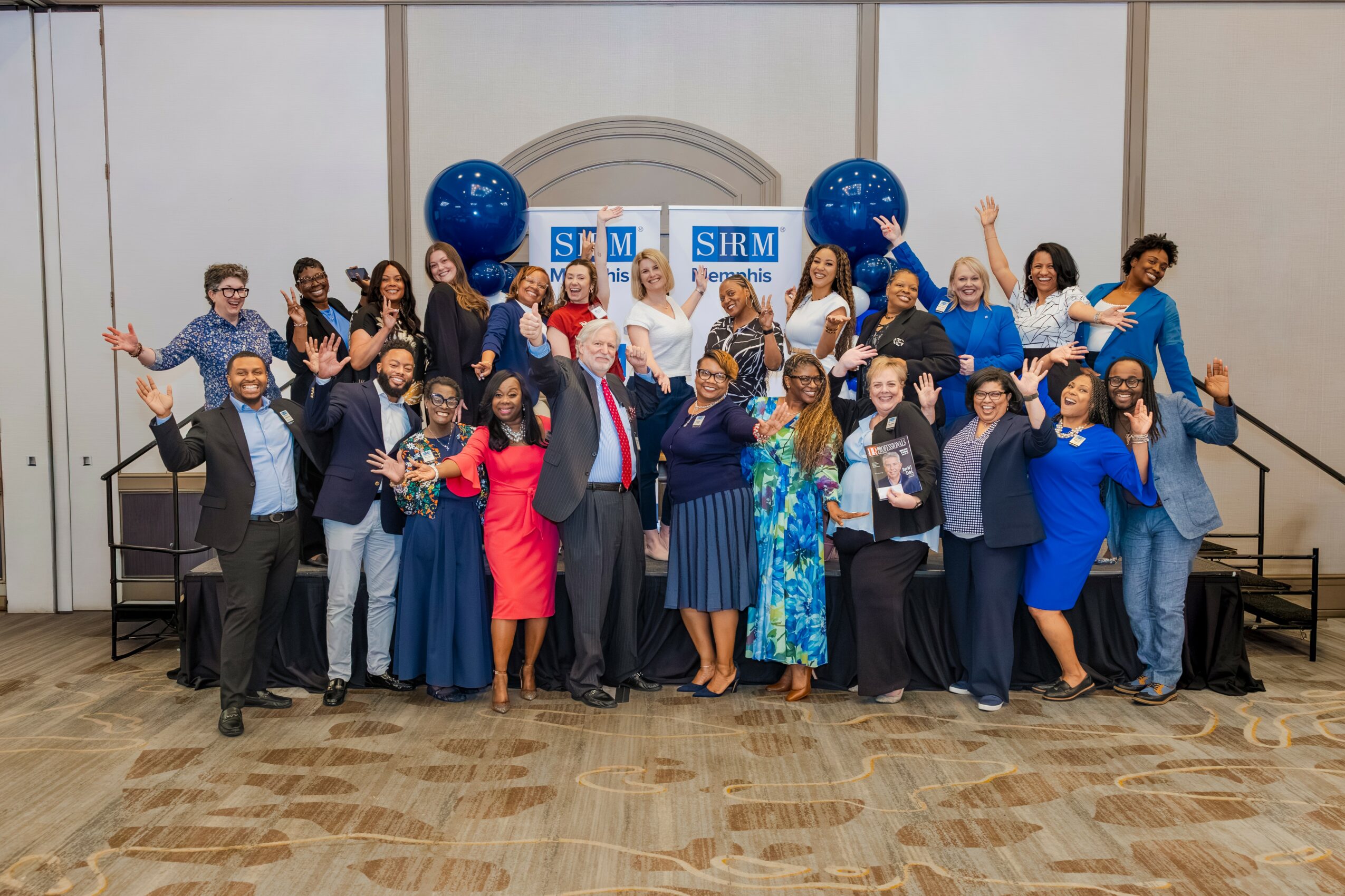 Group of people on stage celebrating, some holding certificates, with blue balloons and a banner in the background.