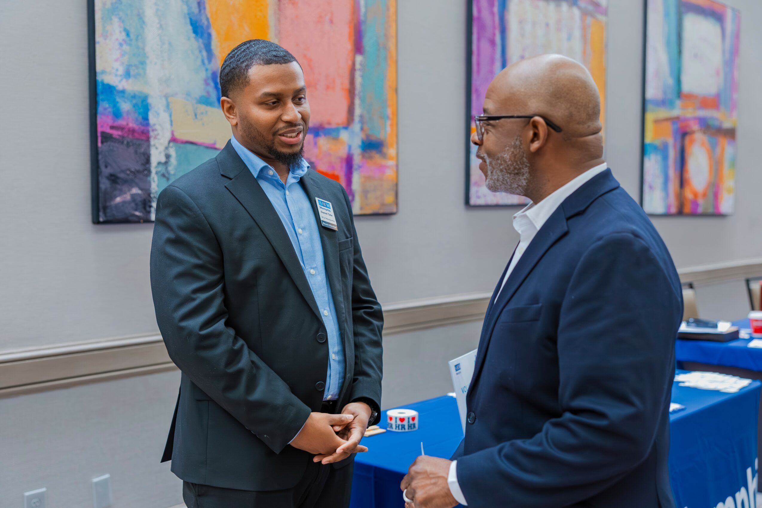 Two men in suits conversing in a room with colorful abstract paintings on the wall.