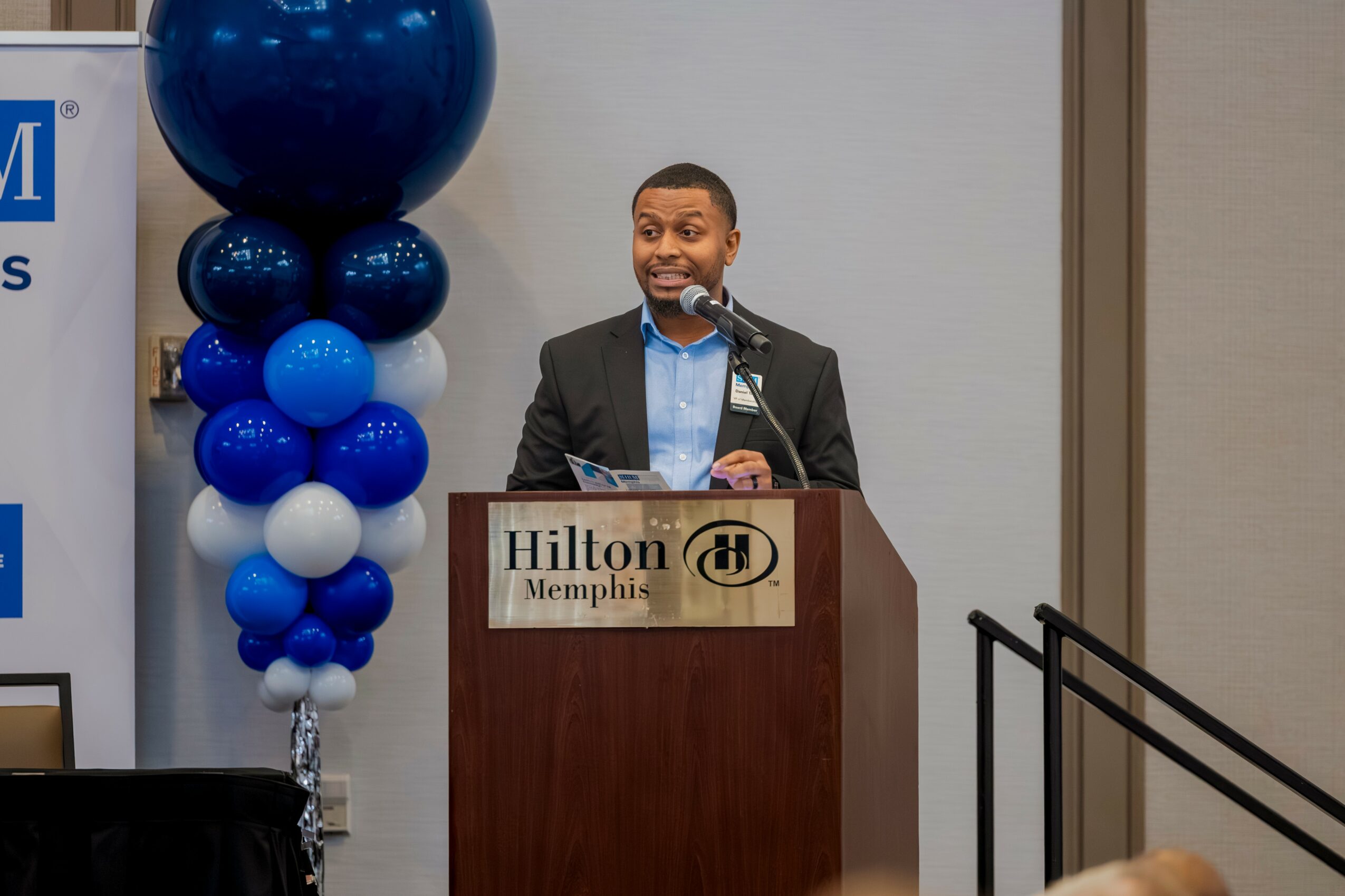 Man in suit speaking at podium with Hilton Memphis sign, decorated with blue balloons, in conference room.
