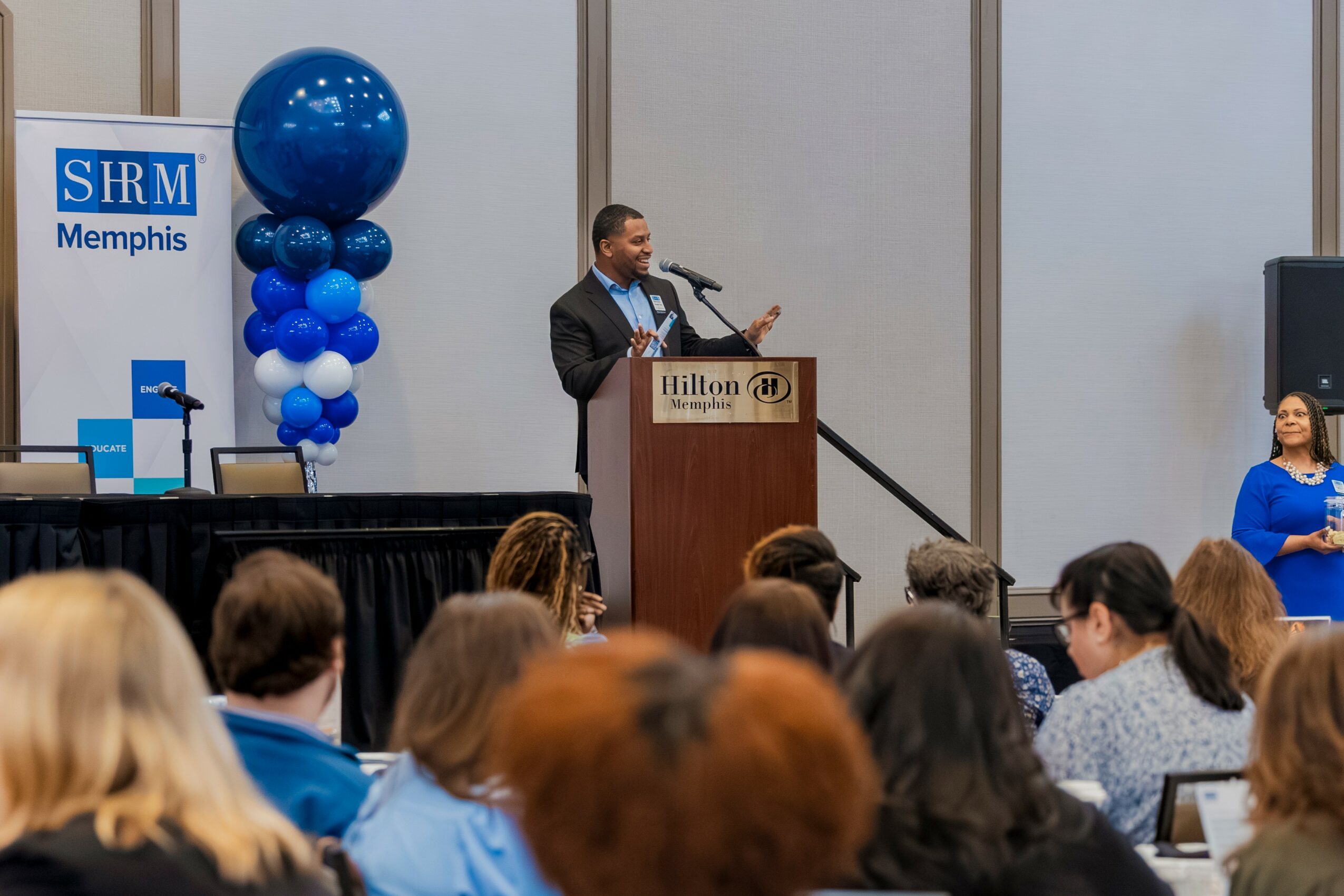 Man speaking at podium during conference, audience seated, blue balloon decoration, Hilton hotel sign, woman in blue dress, SRM Memphis sign.