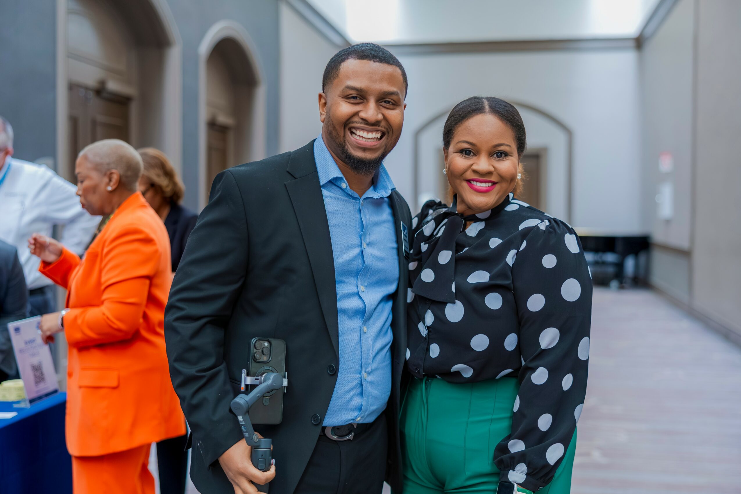 A smiling man and woman pose together indoors, with other people in the background, in a spacious, well-lit area.