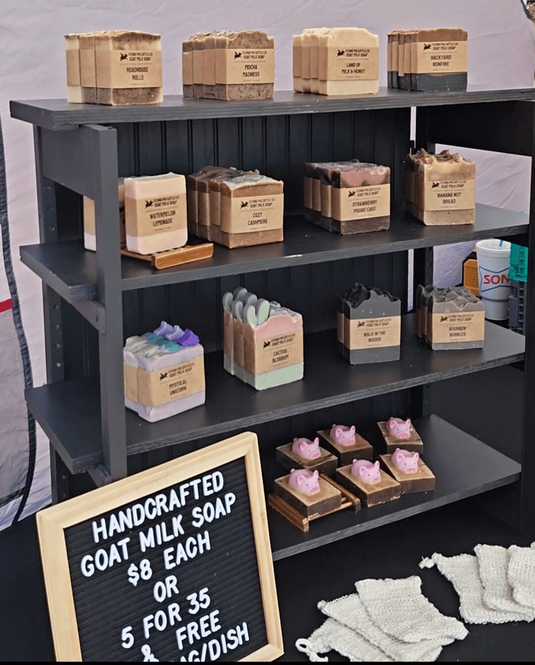 Black shelf with handmade goat milk soap in various shapes and colors, and a chalkboard sign with prices, against a purple wall.