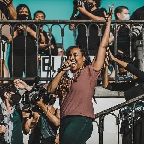 Woman smiling and raising her hand while speaking into a microphone at a protest or rally, surrounded by photographers and people wearing masks.