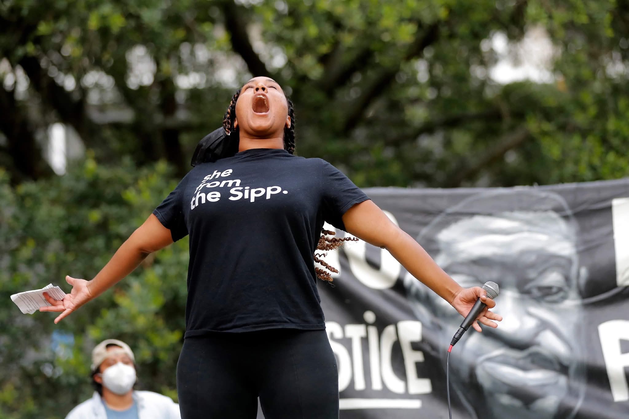 Person with arms outstretched, mouth open, holding a microphone, outdoors with trees and a banner in background.