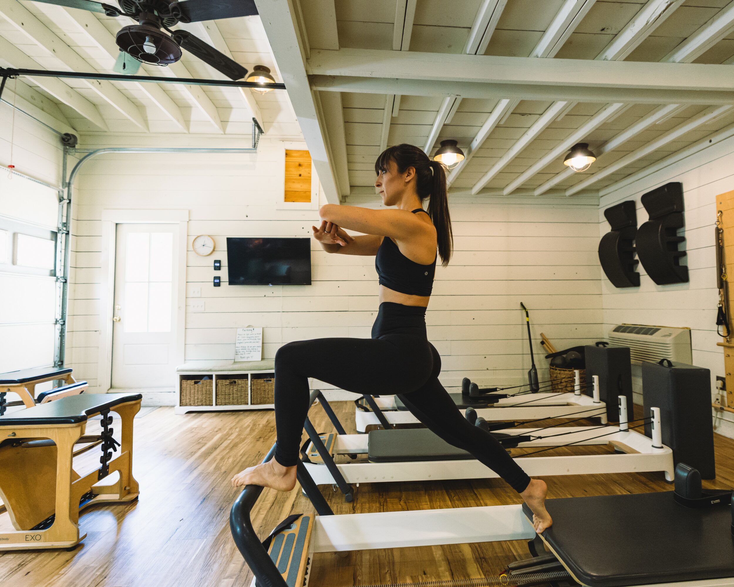 Woman exercising on a treadmill in a gym with wooden floors and white walls, TV mounted on wall, and exercise equipment.