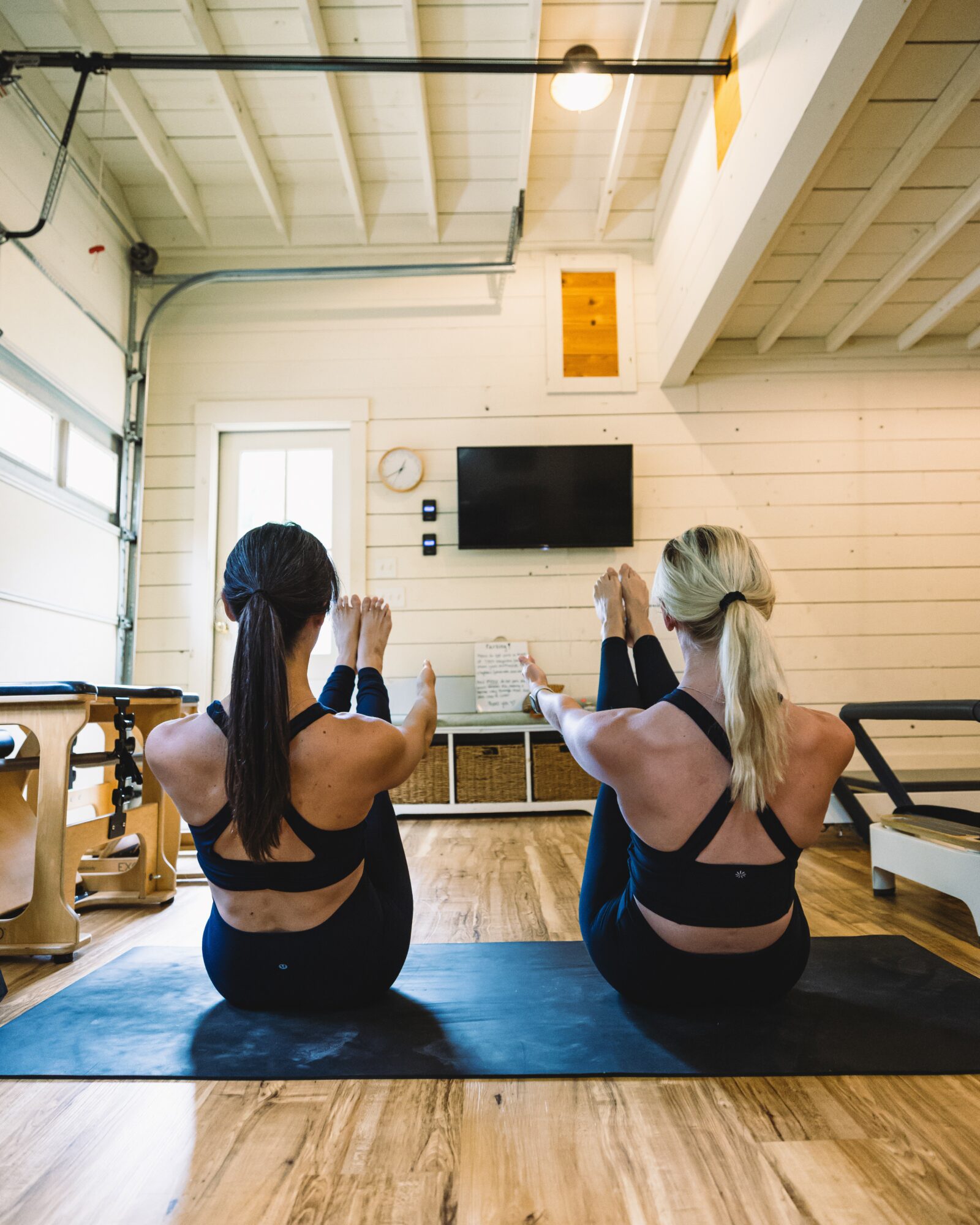 Two women sit on a yoga mat in a bright room, facing a wall with a TV and wooden paneling, practicing yoga.