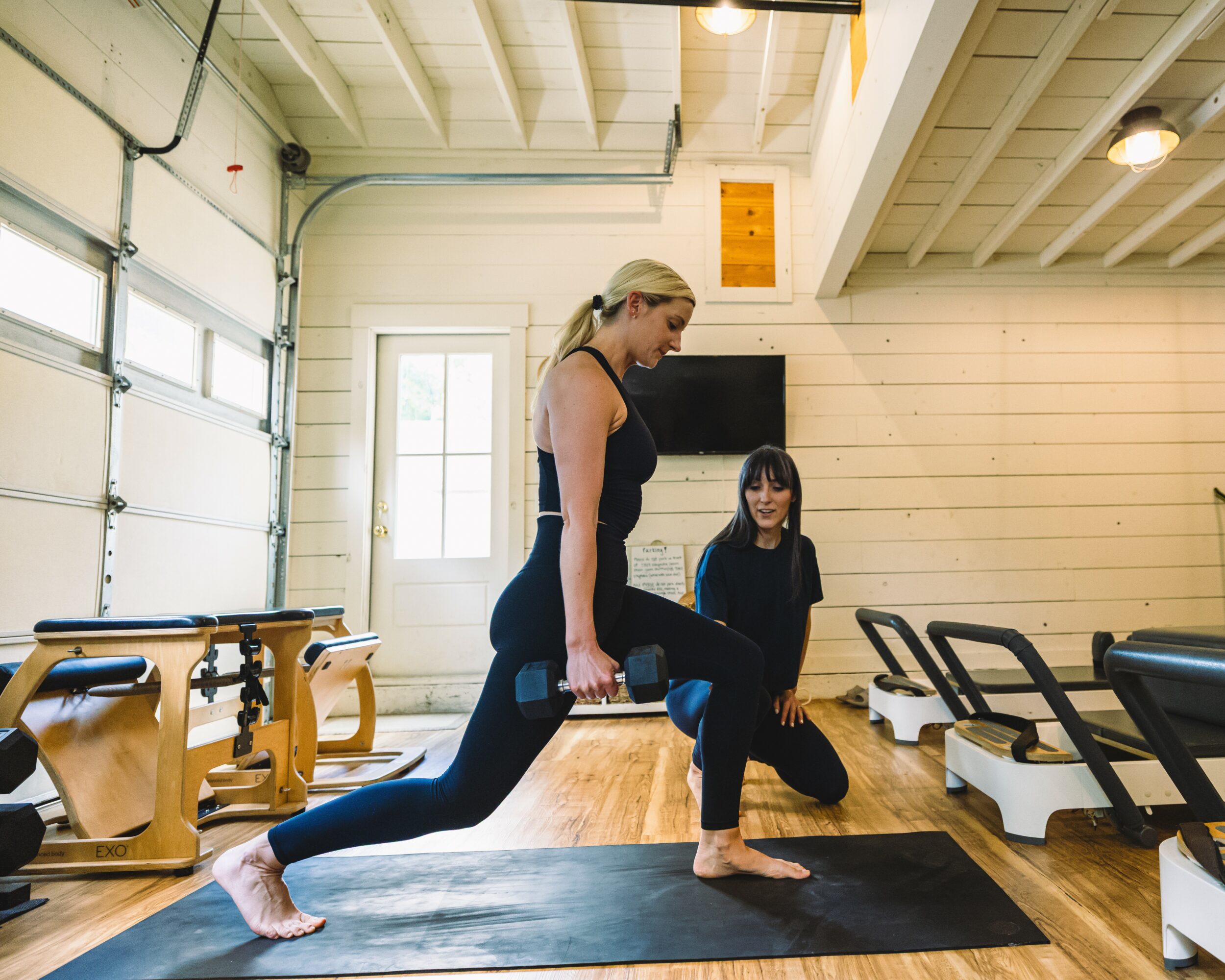 Two women practicing yoga in a gym, one in a lunge pose, the other kneeling, with gym equipment around them.