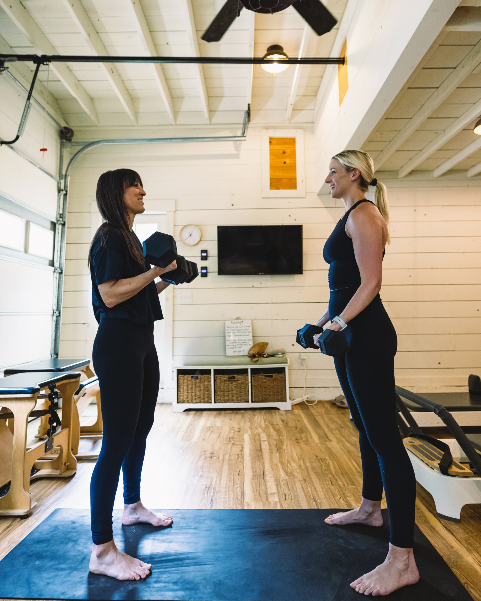 Two women stand facing each other in a gym, holding small weights, with a wooden floor and white walls.