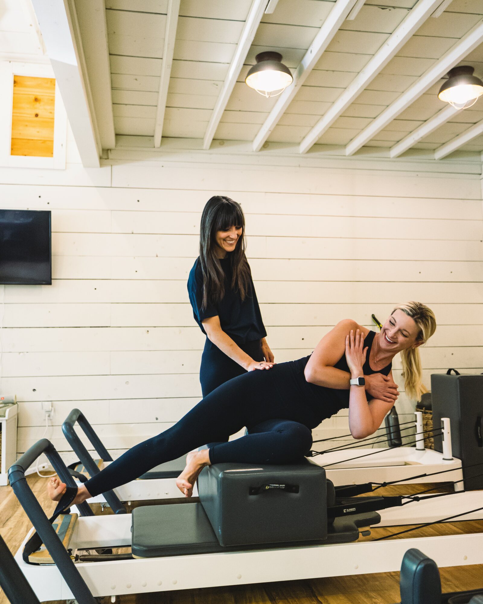 Two women in a fitness studio, one lying on a bench and the other standing beside her, smiling and engaging in exercise.