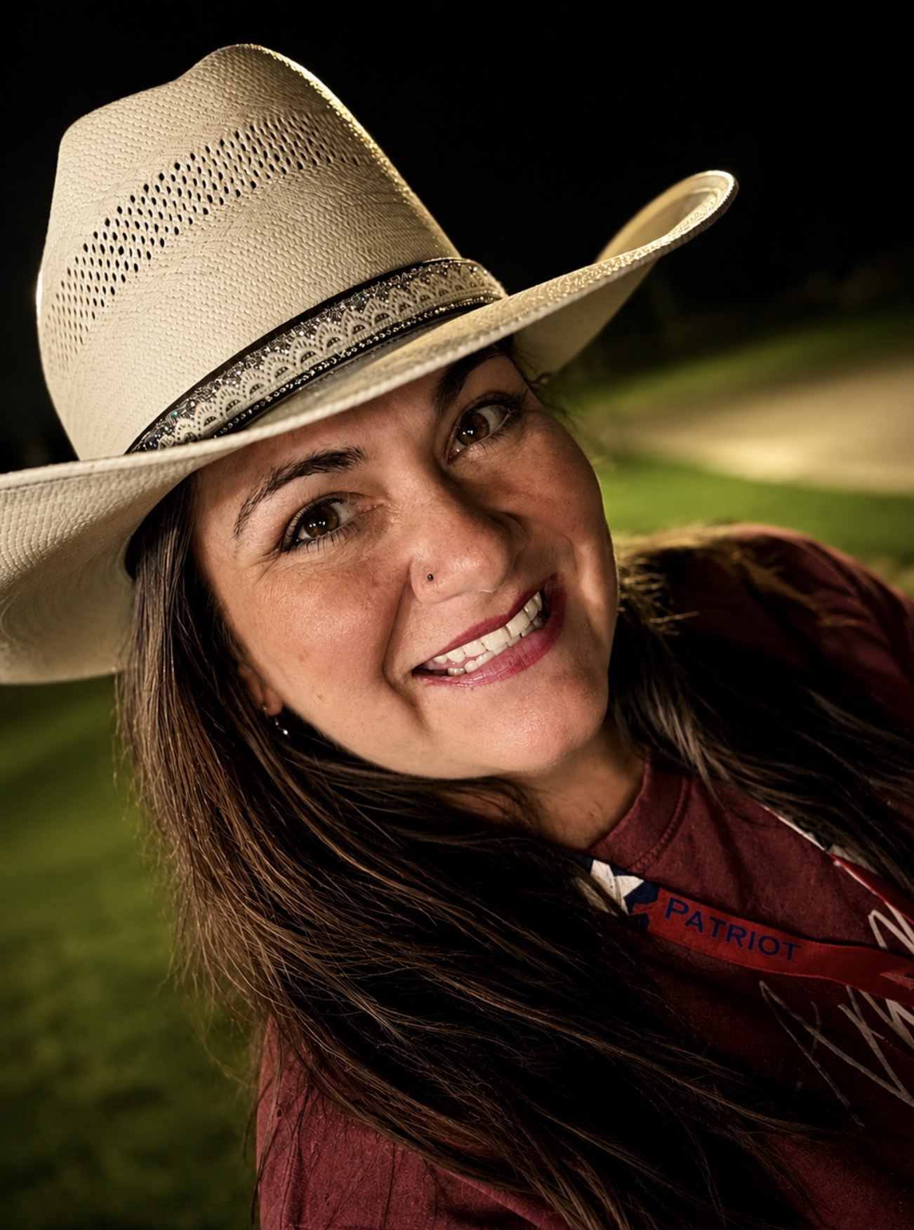 Smiling woman wearing a cowboy hat outdoors at night, with a dark background and blurred greenery.