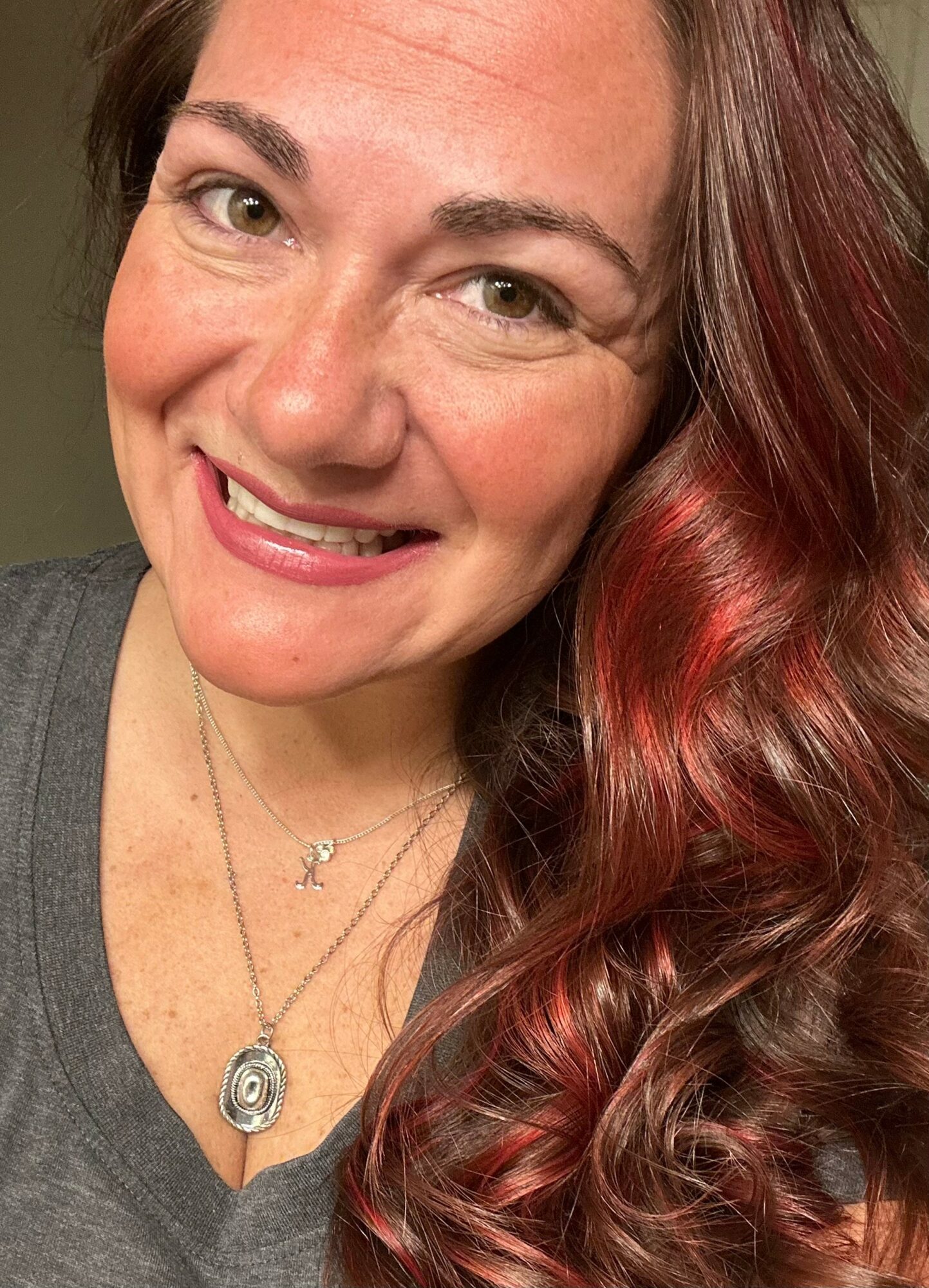 Close-up of smiling woman with long, wavy red hair, wearing a gray top and necklaces.